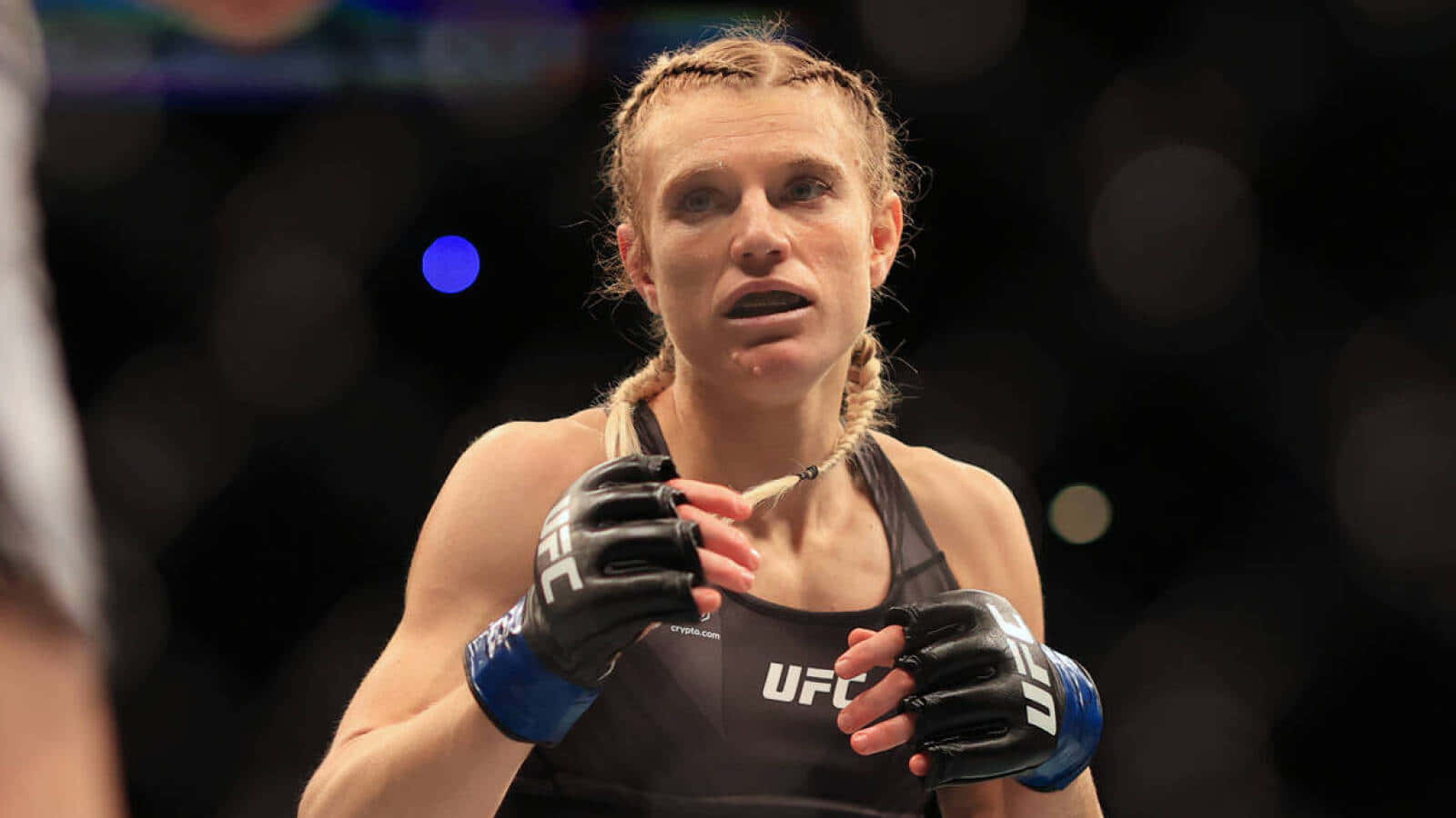 曼恩·菲奧羅特自信展示雙拳 壁紙 — 一位專注的女性MMA拳手，身穿黑色UFC裝備，編織頭髮，在場館燈光下比賽時凝視著。