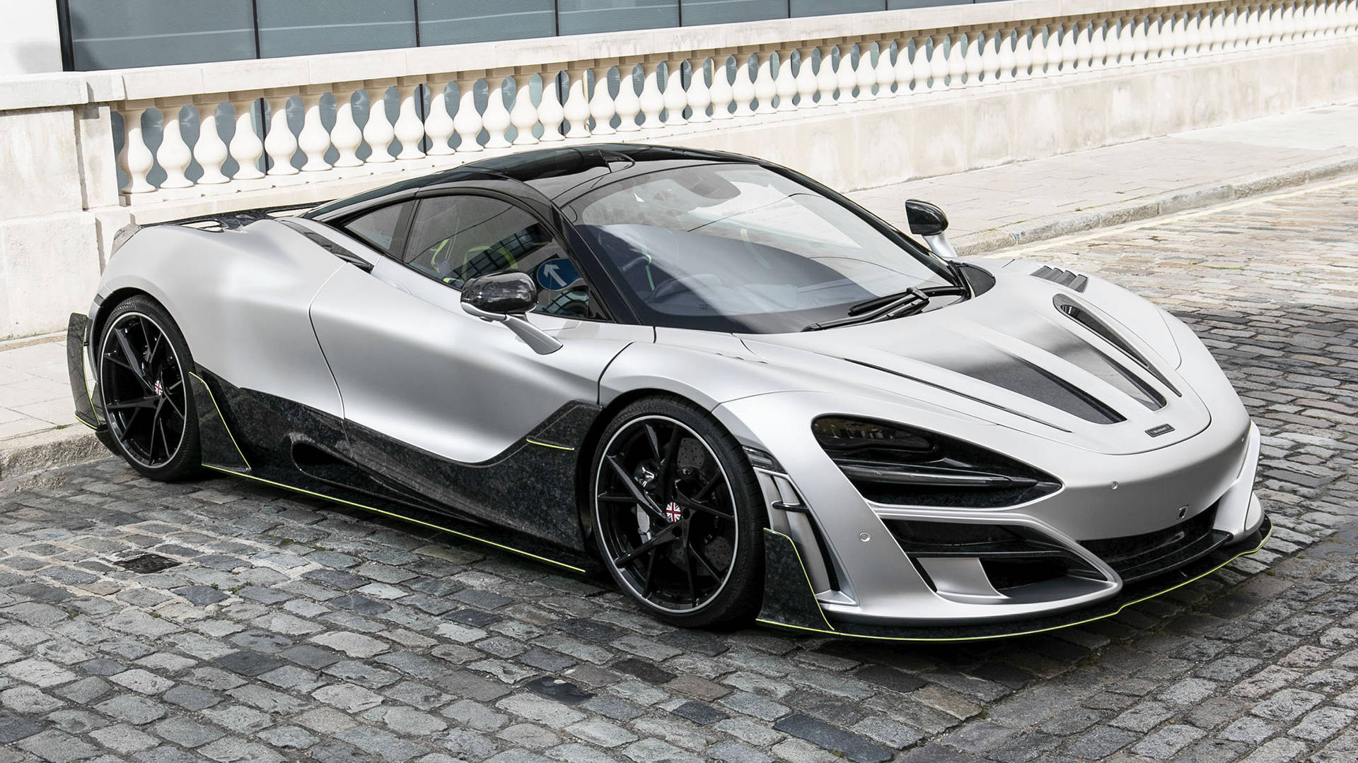 曼索里麥拉倫720S 4K 壁紙 — 一架時尚的銀色McLaren Senna，帶有黑色點綴，停在石欄杆旁邊的鵝卵石街道上。