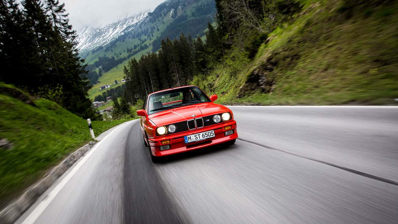 紅色BMW M3在山路上飛馳，有綠色森林和雪山。