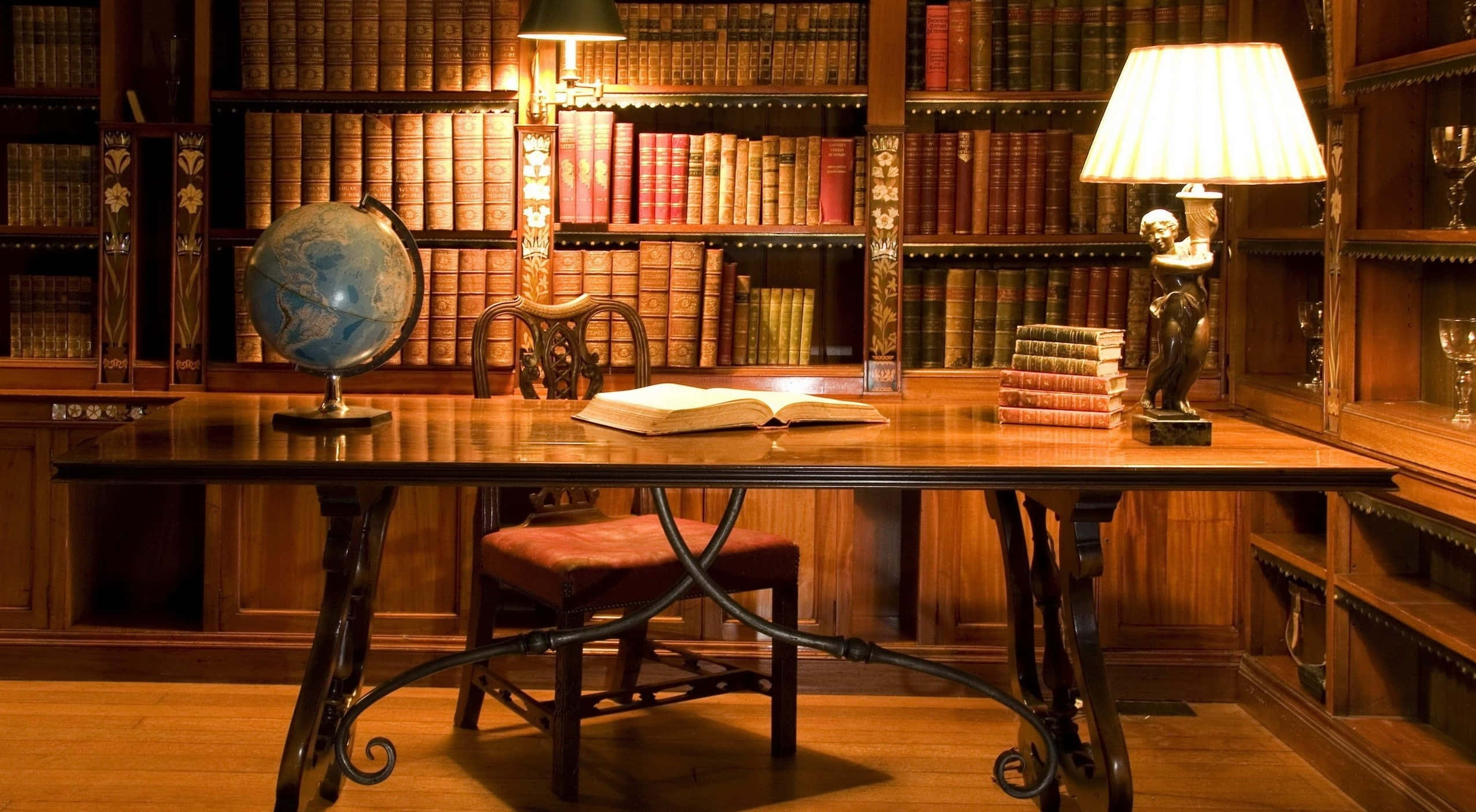 最佳家庭圖書館桌面背景 壁紙 — 一張溫暖照明的木製書桌，上面有打開的書本、地球儀和燈，周圍是高聳的書架。