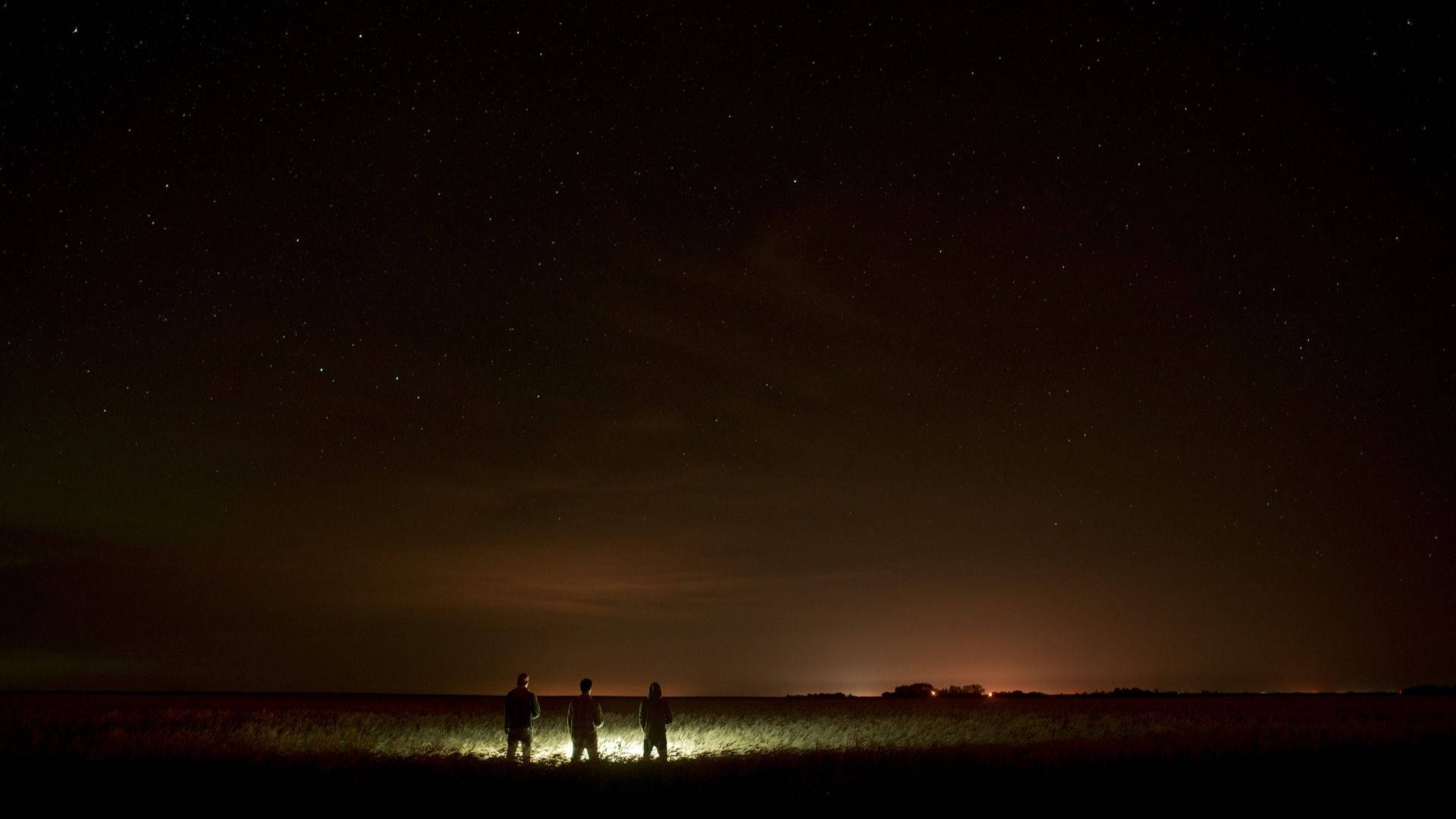 最佳暗夜田野 壁紙 — 三個剪影人物站在草地上，頭上是廣闊的星空，地平線遠處有橙色的光芒。