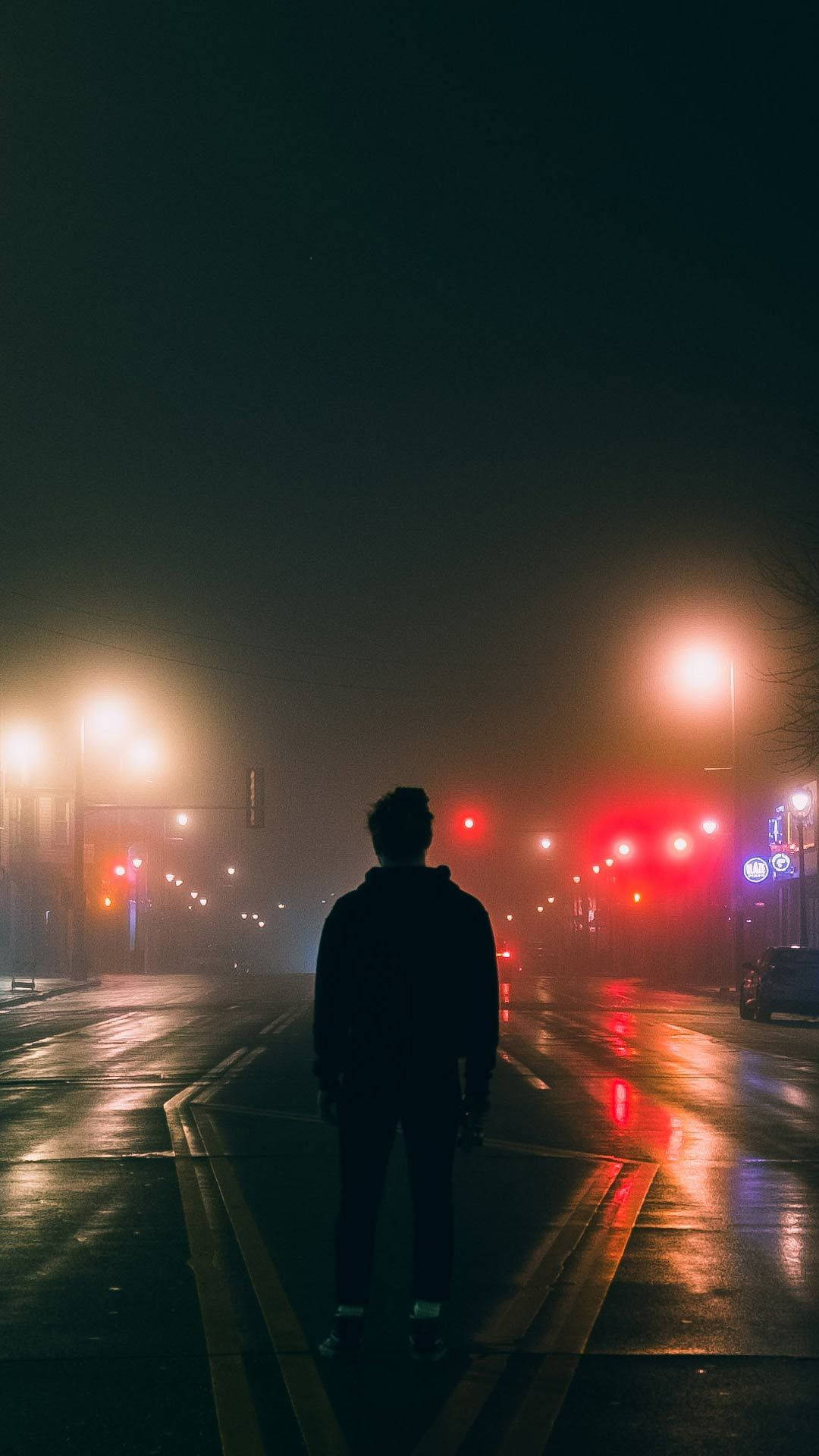 最佳暗夜跑道 壁紙 — 一個孤獨的身影站在霧氣瀰漫的潮濕街道中央，夜晚被發光的街燈和紅色交通信號燈照亮。