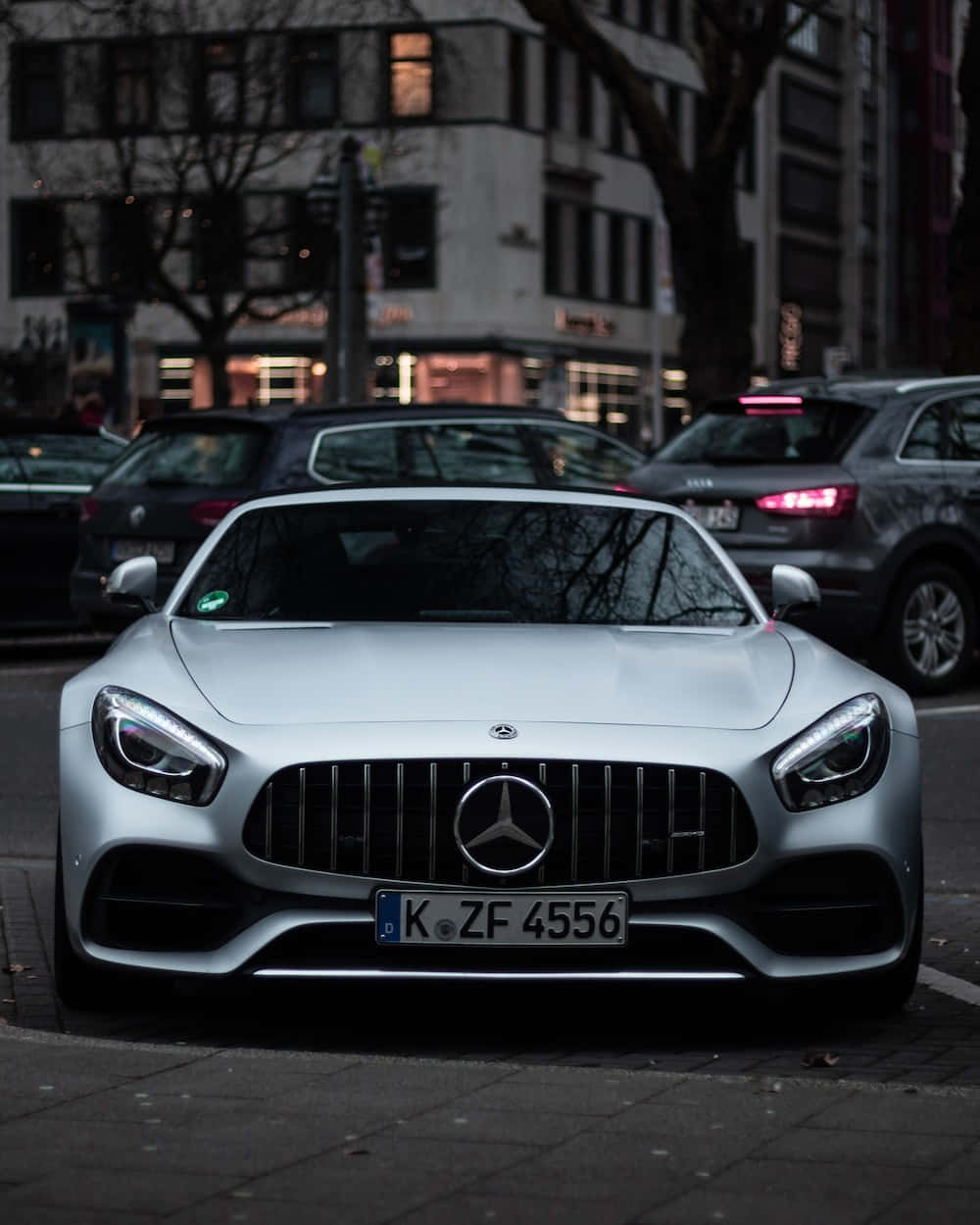 最佳梅賽德斯背景：霧面淺灰AMG GT 壁紙 — 一輛時尚的銀色 Mercedes-AMG GT 在黃昏時分停泊在城市街道上，車頭燈閃爍，背景是都市建築。