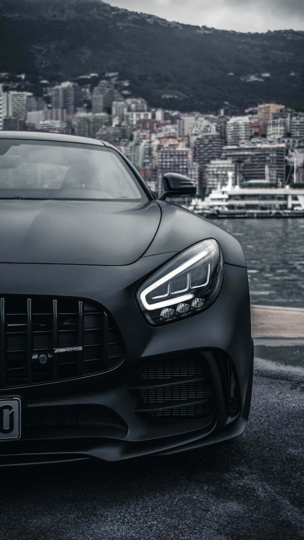 最佳梅賽德斯背景：霧面黑色AMG GT 壁紙 — 一架啞黑色AMG跑車停泊在海濱，背景是城市天際線和山丘。