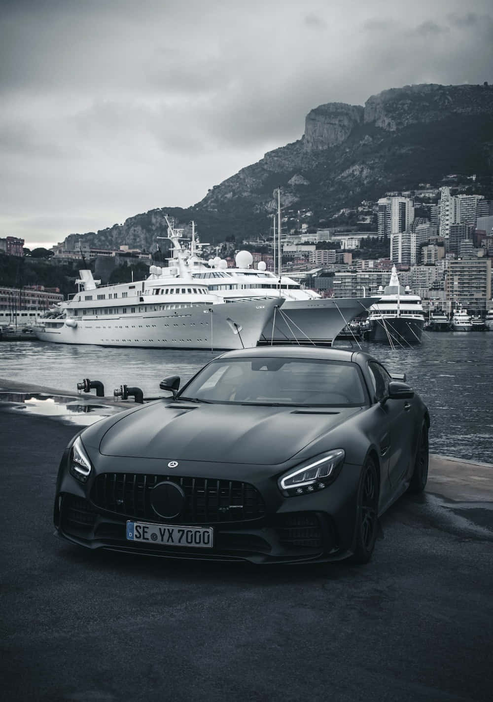 最佳梅賽德斯背景：黑色AMG GT停靠碼頭旁 壁紙 — 一架啞黑色跑車停泊在港口碼頭，背景有豪華遊艇和懸崖城市，天色陰沉。