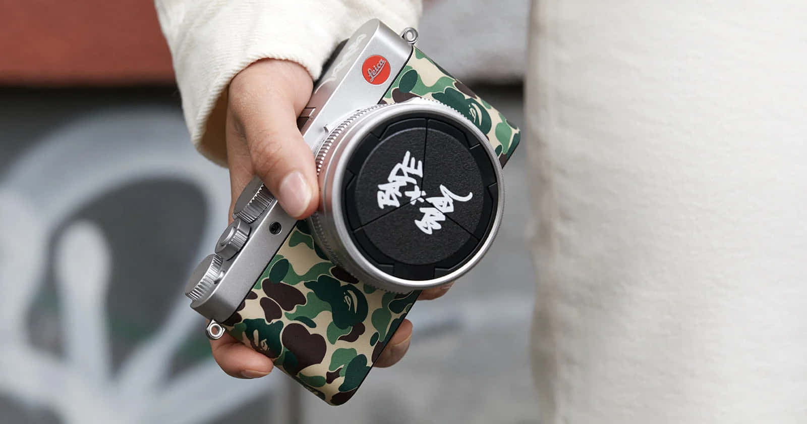 手拿著帶有迷彩機殼和塗鴉鏡頭蓋的銀色 Leica 相機。