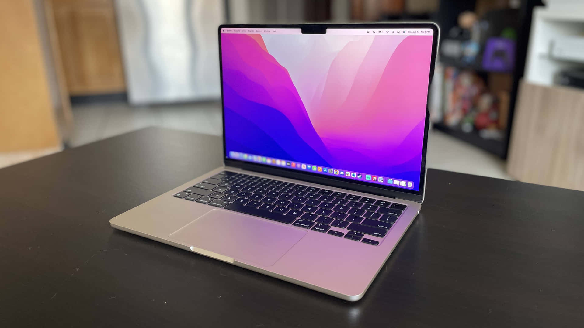 最新款 Macbook 搭配鍵盤與觸控板 壁紙 — 一部銀色手提電腦，顯示著充滿活力的粉紅和紫色抽象桌布，打開放在深色木桌上，房間光線柔和。