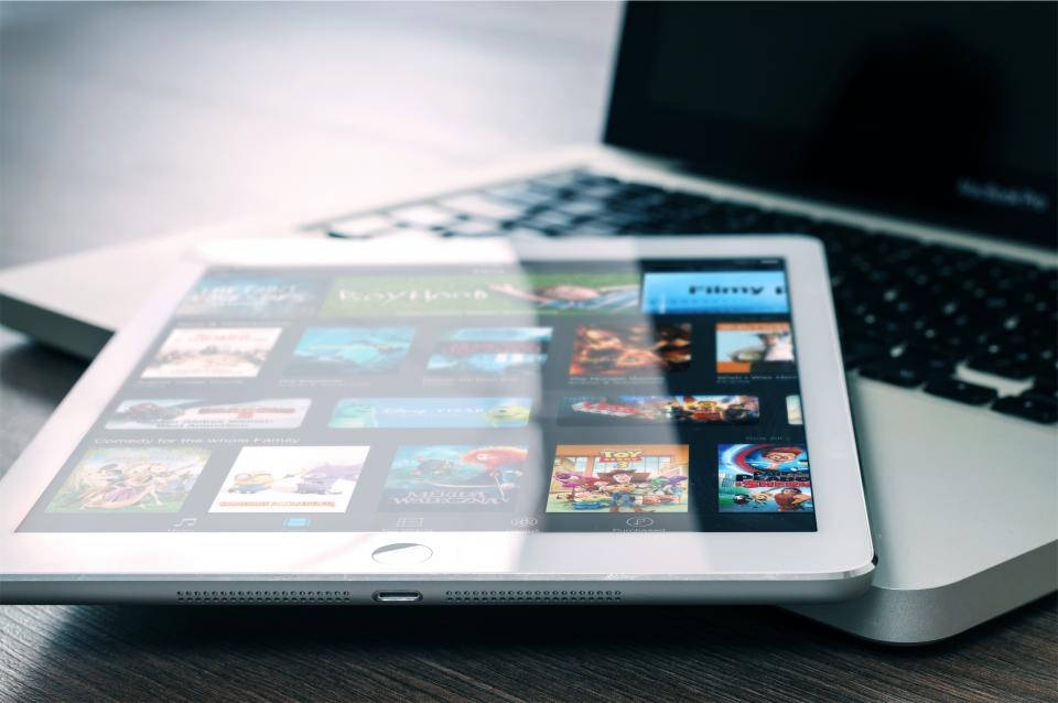 最新款iPad Pro白色背景展示 壁紙 — 一部銀色平板電腦顯示色彩繽紛的電影縮圖，放在木桌上，旁邊有手提電腦鍵盤。