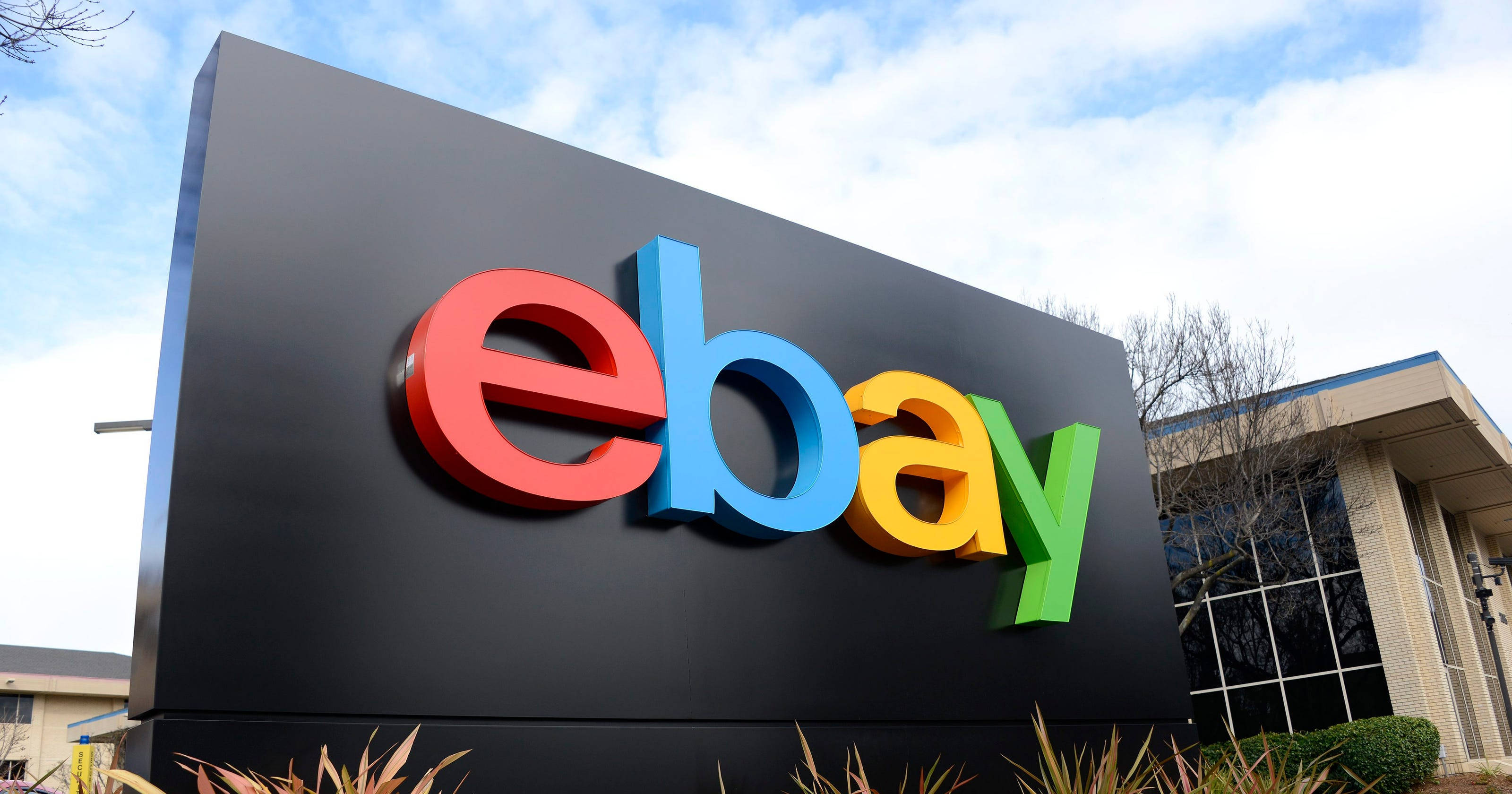 最新版 eBay 標誌招牌 壁紙 — 一個大型彩色eBay標誌牌在部分多雲的天空下醒目地矗立，附近有現代建築和乾燥植物。