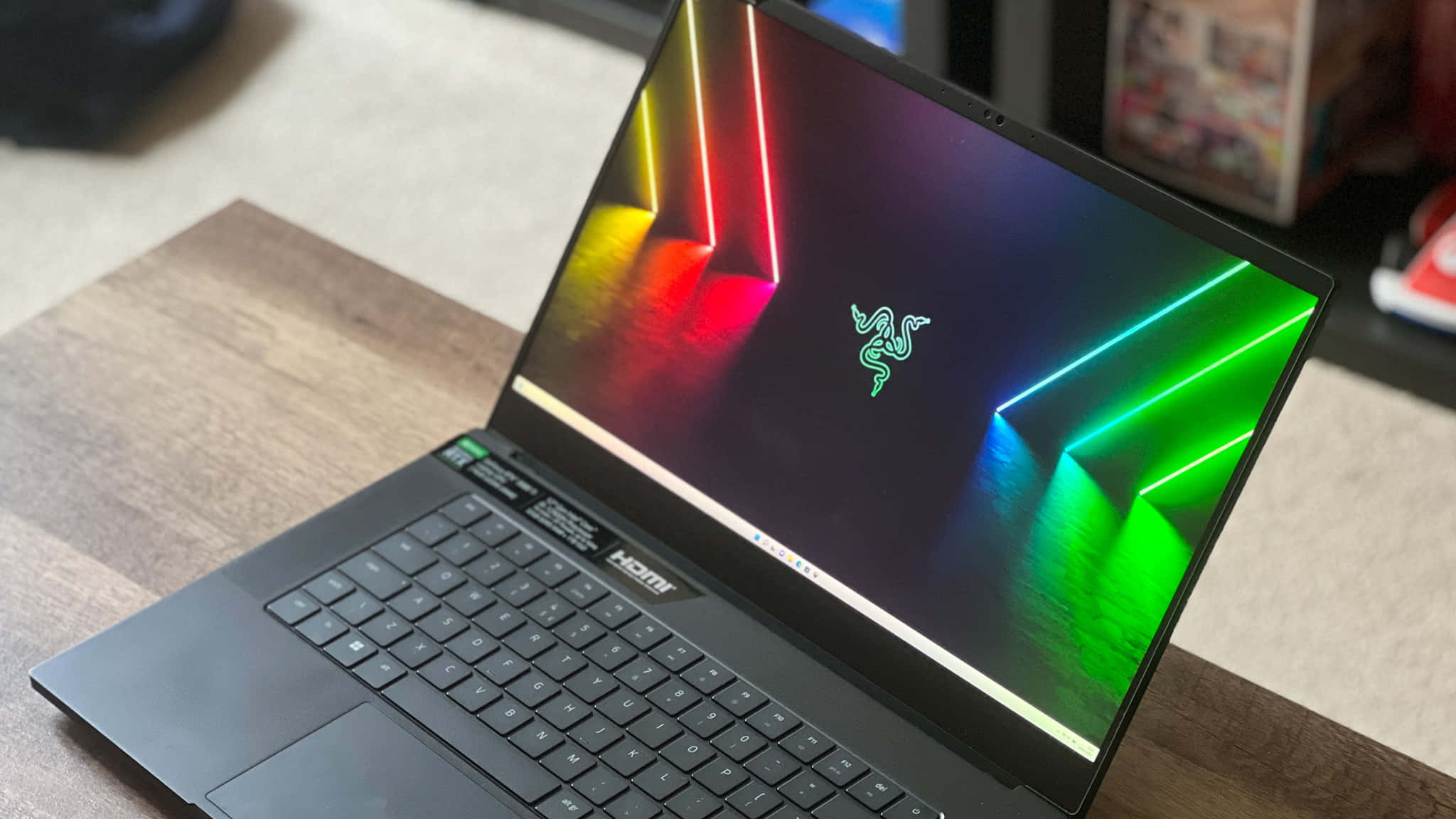 最新筆電，為您的工作流程注入動力 壁紙 — 一部時尚的黑色 Razer 筆記型電腦在木桌上打開，顯示充滿活力的霓虹燈牆紙，帶有發光的綠色和紅色幾何線條。