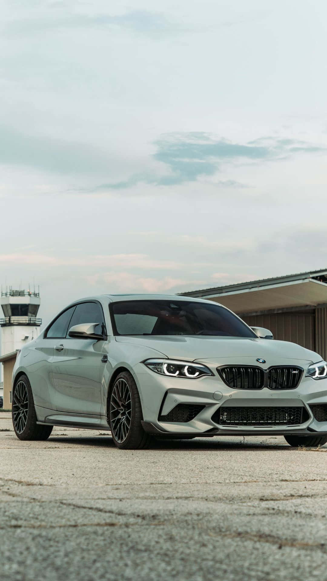 最新豪華駕駛體驗 — BMW安卓整合 壁紙 — 一架時尚的銀色BMW M2雙門跑車停泊在裂開的路面上，背景是機場控制塔，天空多雲。