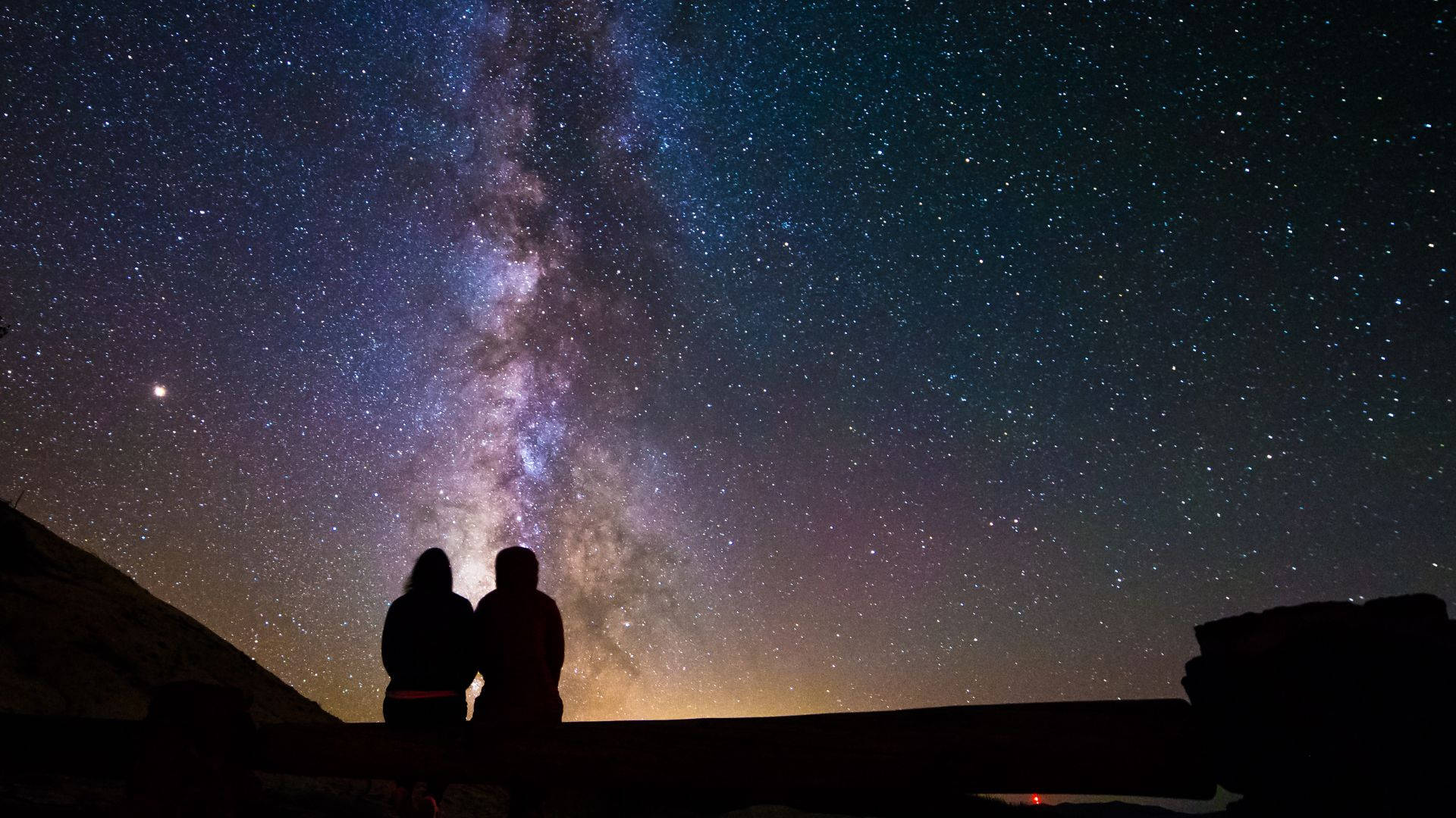 最美的深邃星空夜景 壁紙 — 兩個剪影人物坐在一起，仰望橫跨繁星夜空、充滿活力的銀河系。