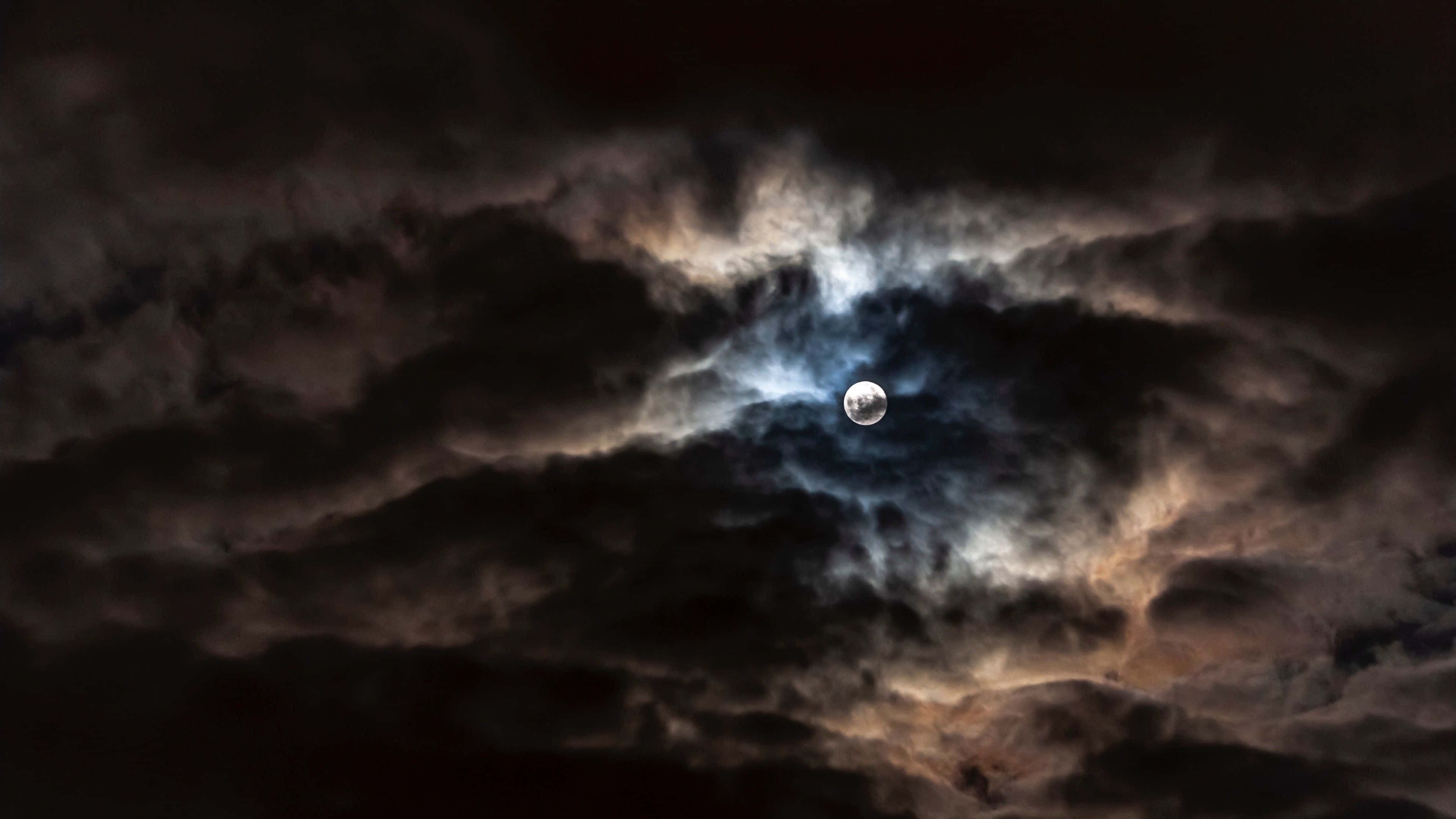 月亮穿過雲層 壁紙 — 一輪明亮的月亮穿透戲劇性的烏雲，散發著微妙的藍色和橙色光芒。