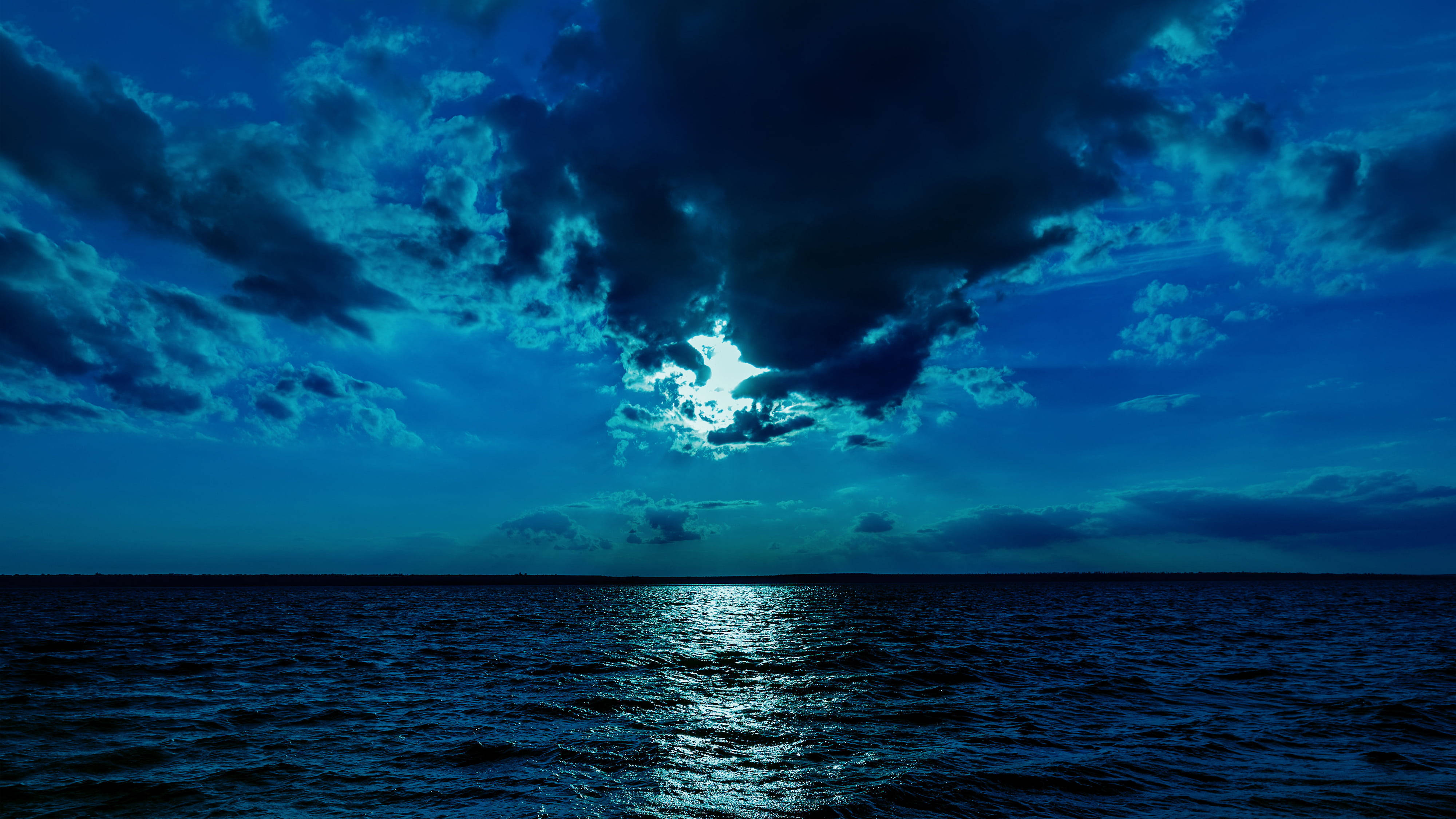 月光下的水域 4K 壁紙 — 月光穿透戲劇性的雲層，在夜晚廣闊的深藍色海洋上投射出閃爍的倒影。
