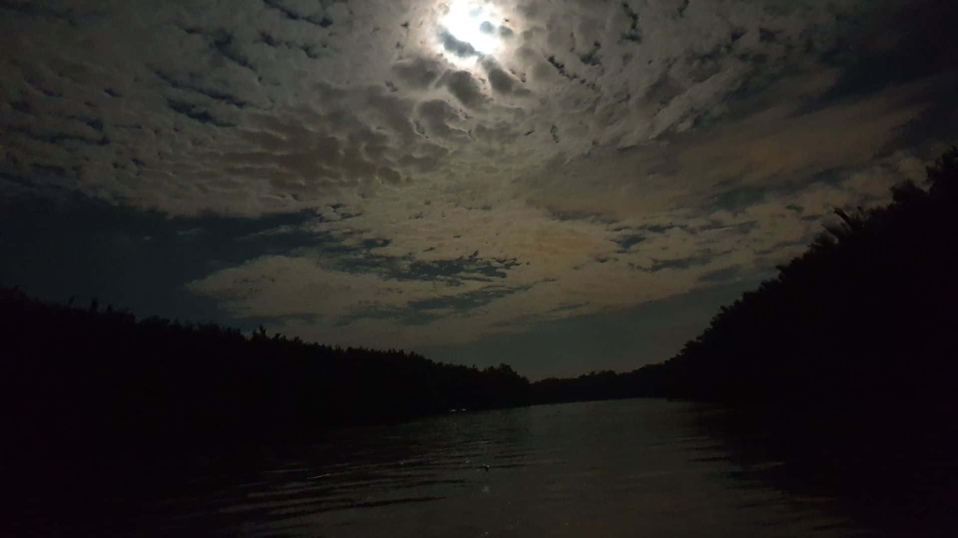Dark Night 桌布 壁紙 — 月光穿透有紋理的雲層，照耀在兩旁被茂密森林剪影環繞的黑暗河流上。
