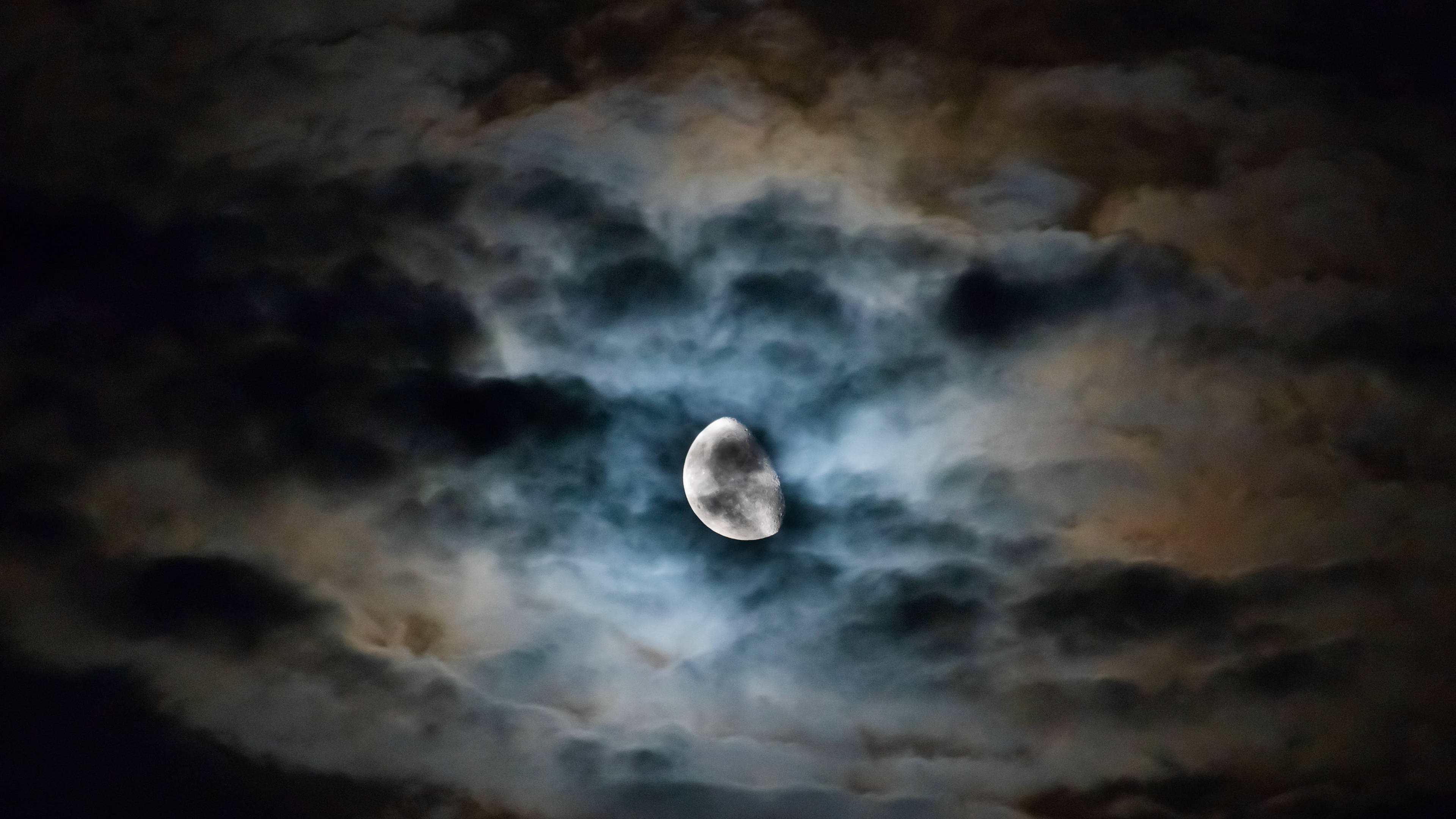 月光穿透烏雲 4K 壁紙 — 一彎新月穿過旋轉的烏雲，帶有藍色和橙色的色調，在戲劇性的夜空中發光。