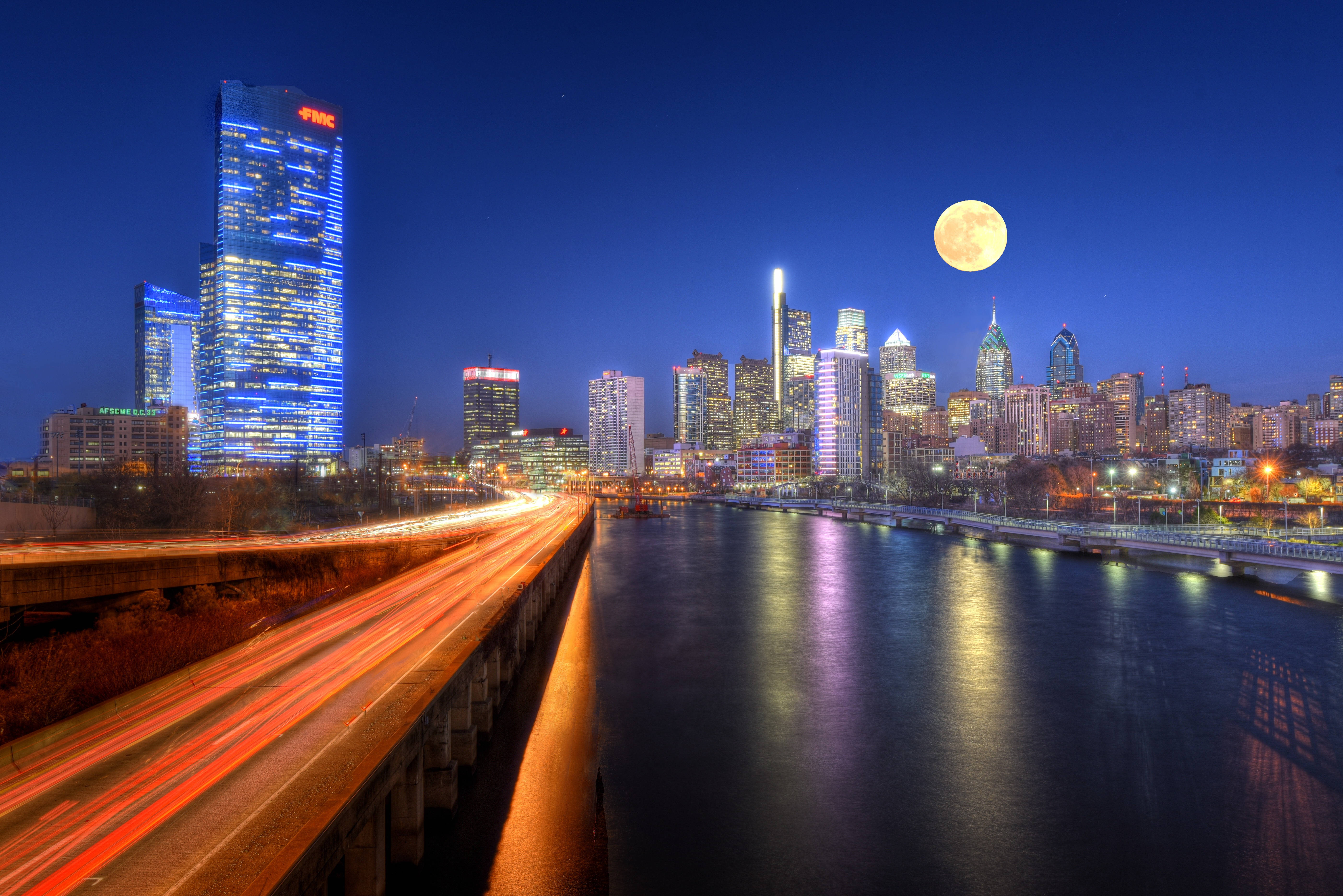 月升費城 壁紙 — 充滿活力的城市夜景，滿月在摩天大樓上方閃耀，繁忙高速公路的光跡在反射的河流旁。