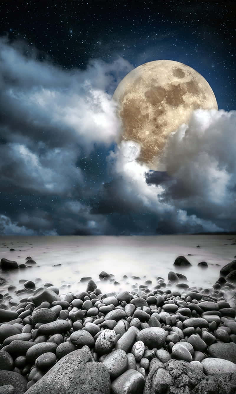 月夜岩石海灘圖 壁紙 — 一個巨大而細節豐富的月亮從戲劇性的雲層中窺視，籠罩在星夜下霧氣瀰漫的岩石海岸線之上。