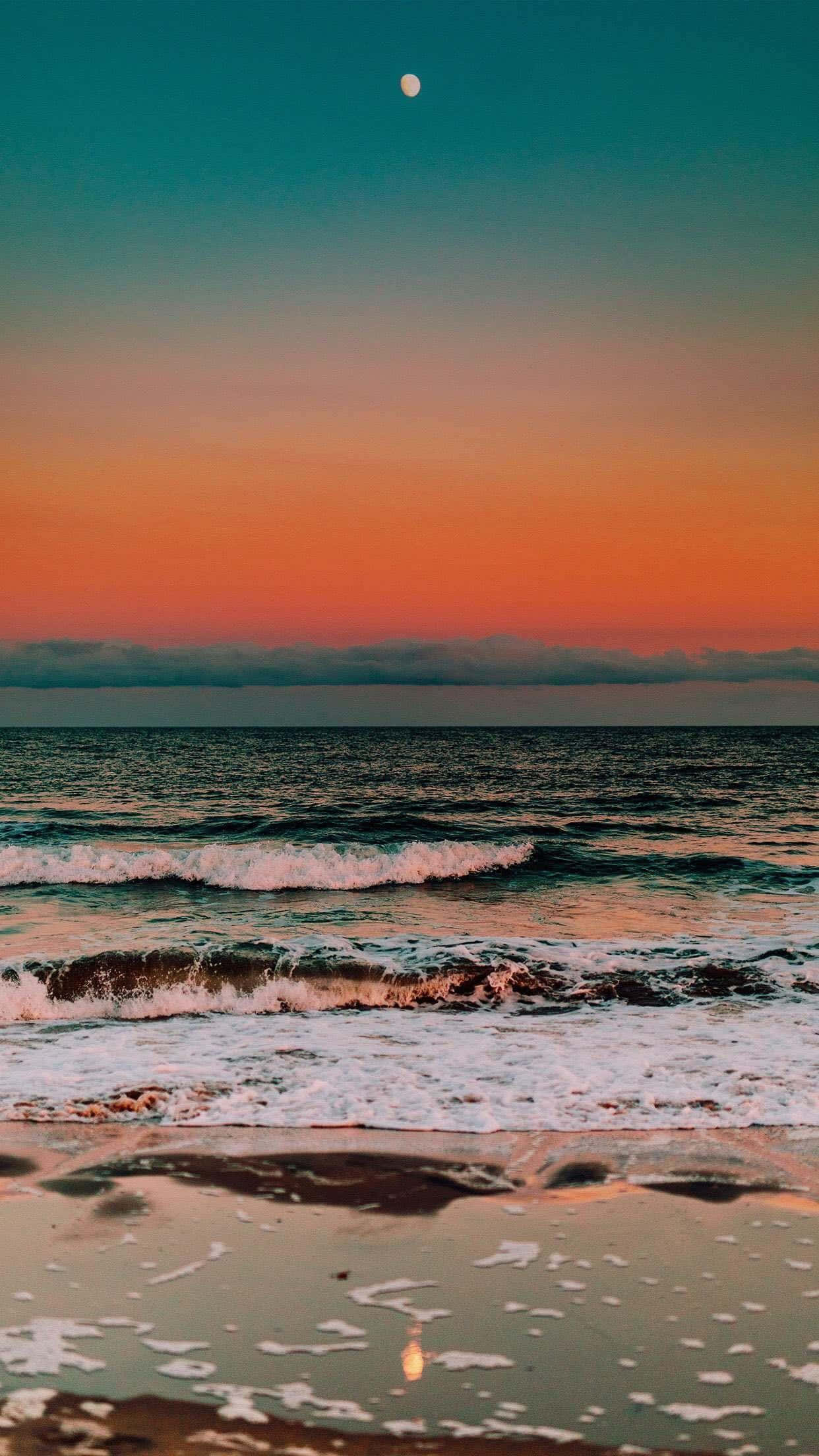 月夜海灘風景圖 壁紙 — 黃昏時分寧靜的海灘，在青綠色、橙色和粉紅色的漸變天空下，溫柔的海浪翻滾而來，月亮清晰可見。