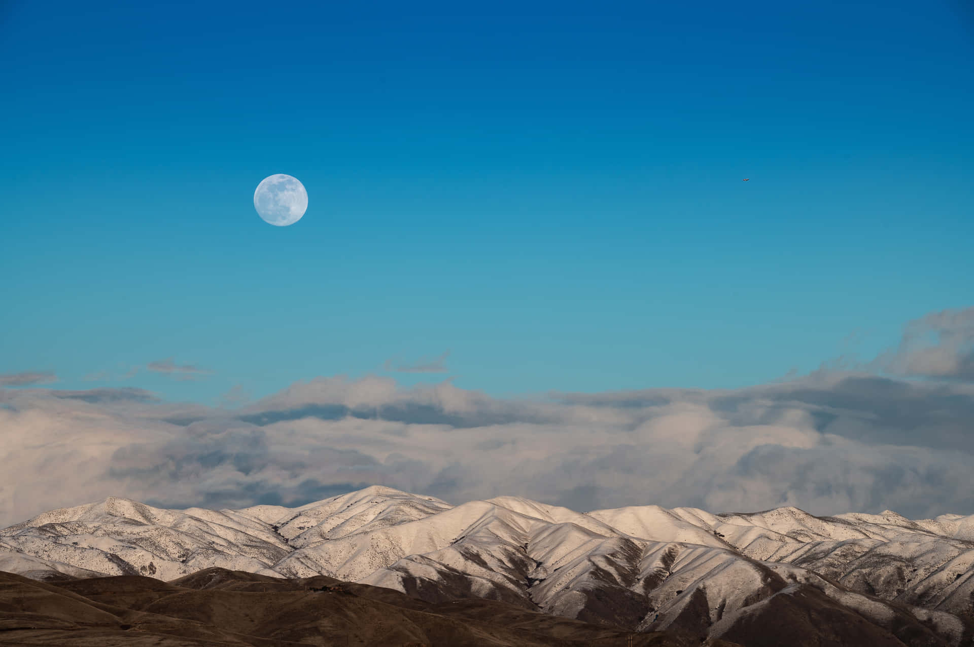 滿月高掛在積雪的山脈和雲層之上，天空清澈湛藍。