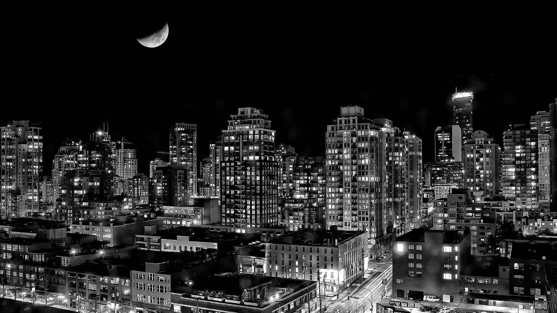 黑白城市天際線夜景，新月在亮燈的建築物上方。