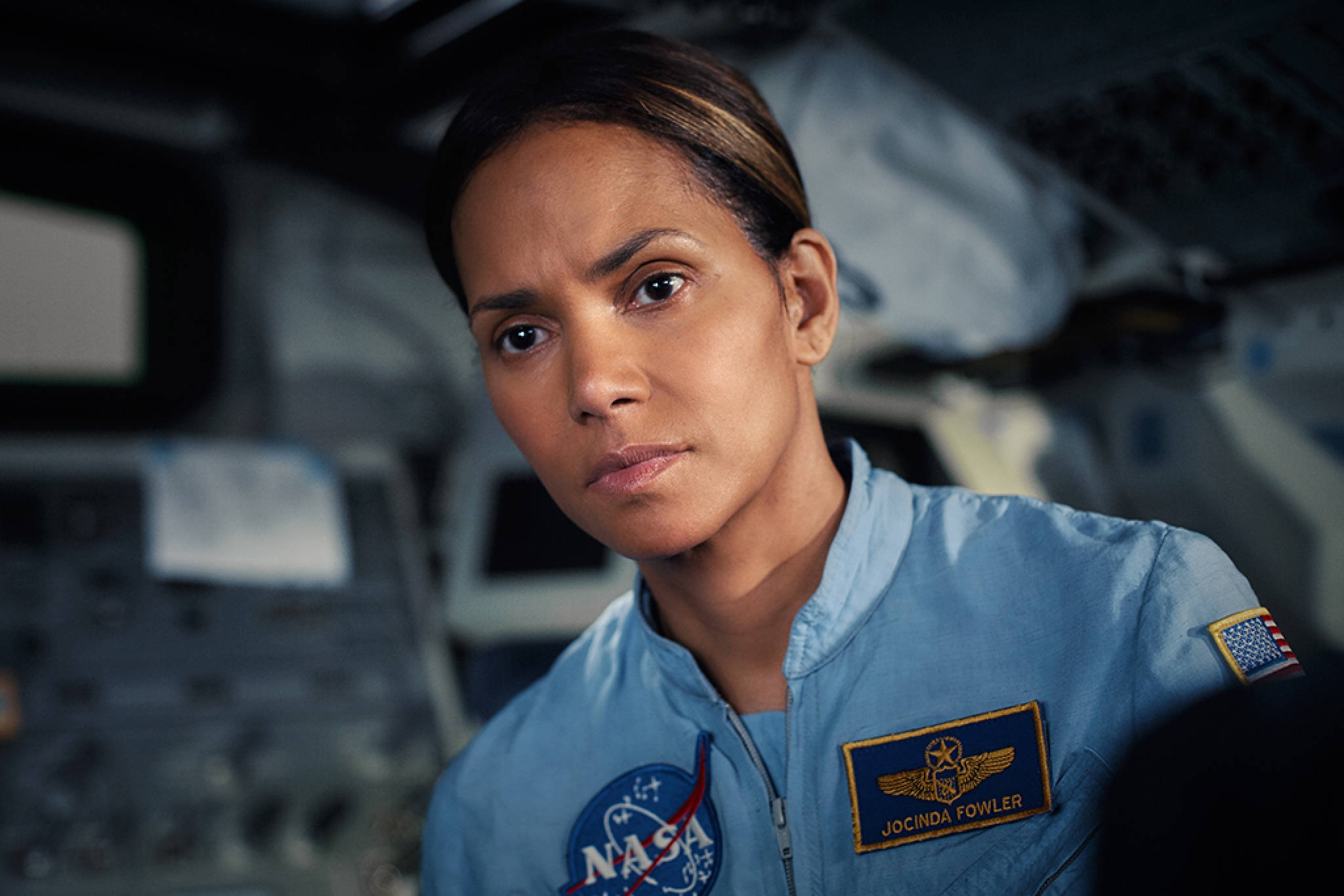 月落Jocinda Fowler 壁紙 — 一位專注的女太空人穿著藍色NASA飛行服，在太空船駕駛艙內凝視著。