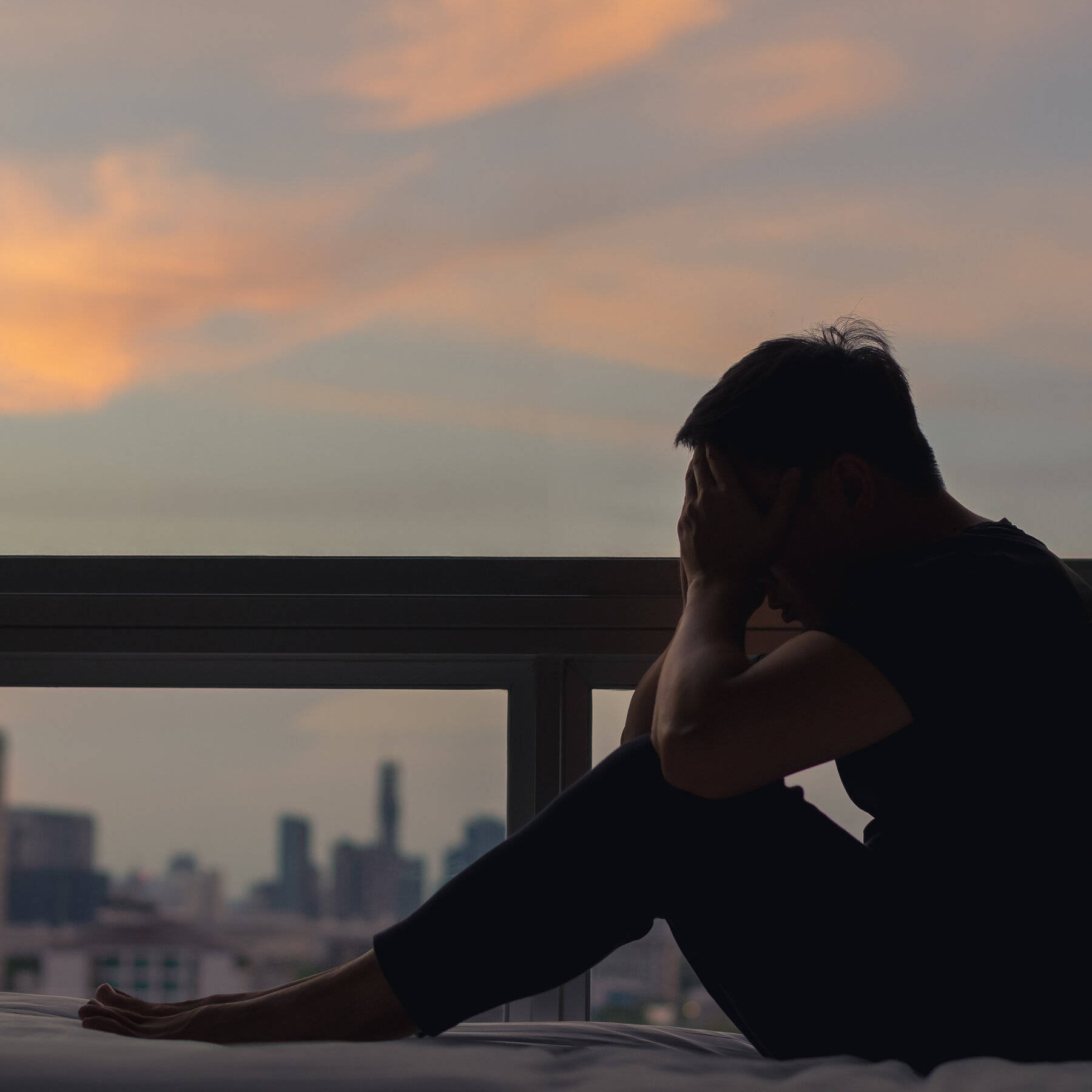 有助表達的媒介 壁紙 — 一個剪影人物坐在陽台的床上，夕陽西下，雙手掩面，俯瞰朦朧的城市天際線。