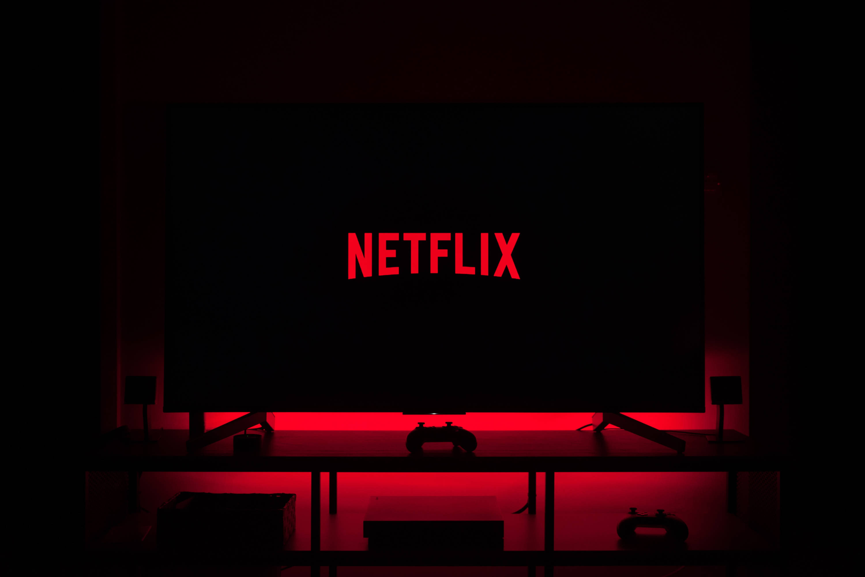 朋友享受 Netflix 影院之夜 壁紙 — 一個黑暗的房間，被顯示Netflix標誌的電視屏幕發出的紅光照亮，下方支架上有遊戲控制器。