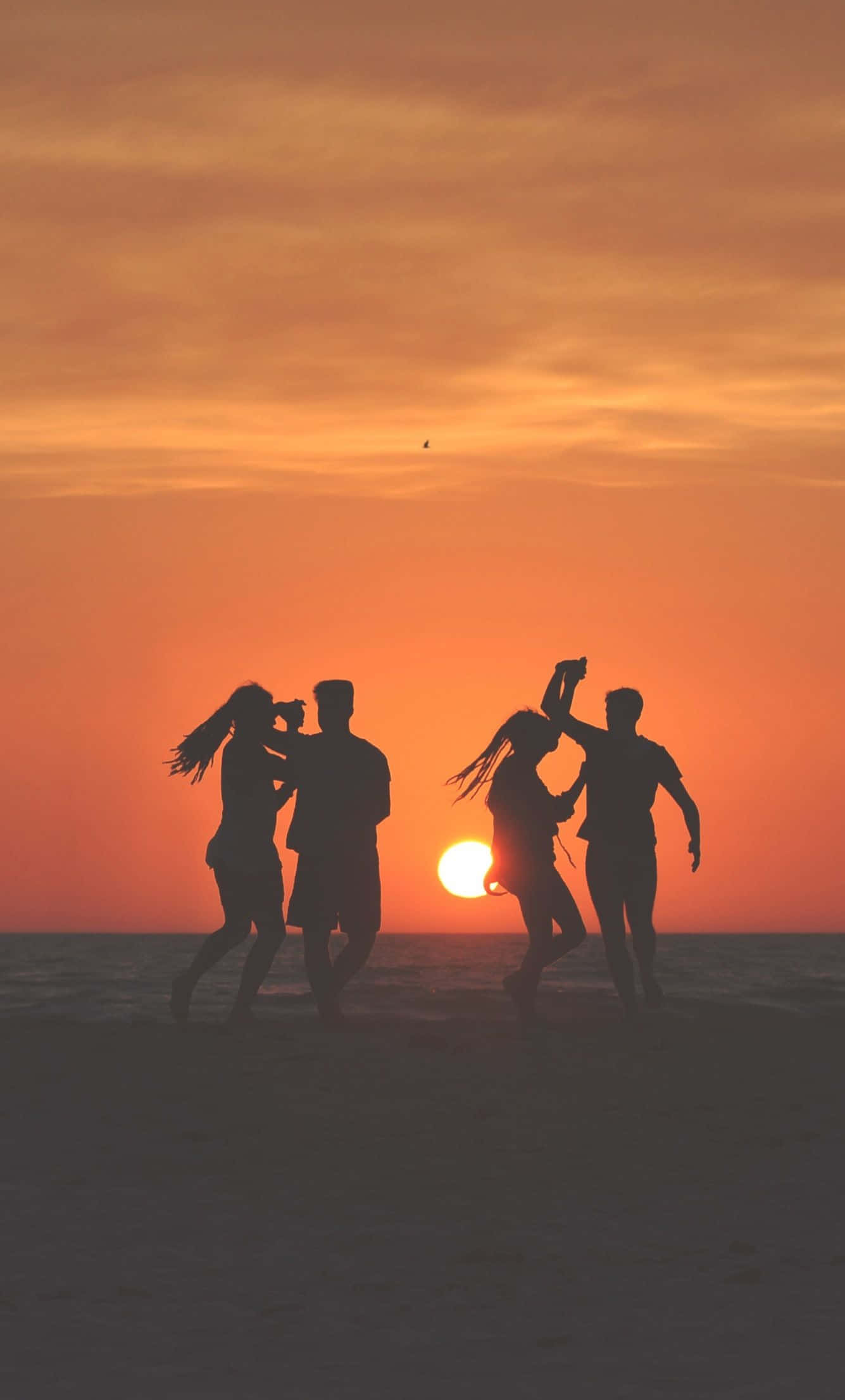 朋友們在海灘熱舞派對歡樂起舞 壁紙 — 四個剪影朋友在沙灘上歡快地跳舞，夕陽呈現火熱的橙色和粉紅色調。