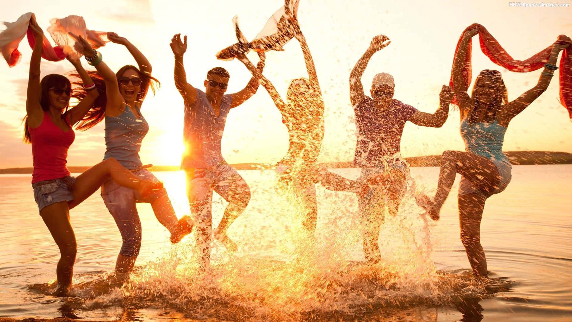 Beach Dance Party 桌布 壁紙 — 朋友們在金色夕陽的水中歡樂地潑水，同時跳舞並揮舞彩色圍巾。