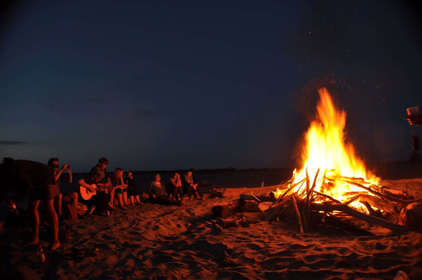 Beach Camping 桌布 壁紙 — 朋友們在黃昏時分聚集在熊熊燃燒的海灘篝火旁，溫暖的橙色光芒灑在沙灘和剪影上。
