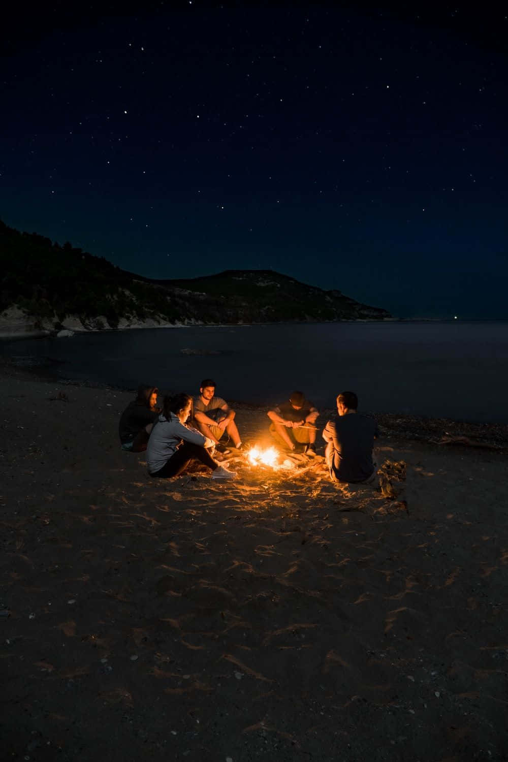 Beach Bonfire 桌布 壁紙 — 朋友們聚集在星光閃爍的海灘上，圍著發光的營火，天空漆黑，遠處有山丘。