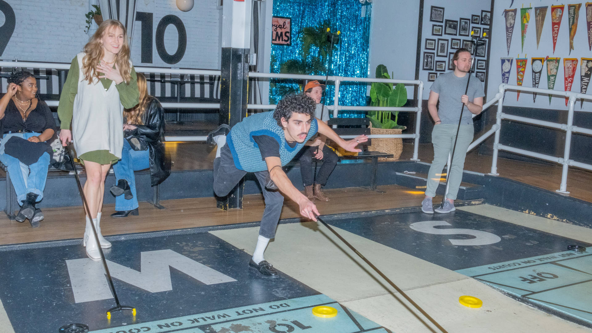 朋友在布魯克林皇家棕櫚 Shuffleboard 俱樂部玩遊戲 壁紙 — 一名身穿藍色背心的年輕男子向前傾身，將冰球推過沙狐球場地，其他人在熱鬧的室內遊戲廳觀看。