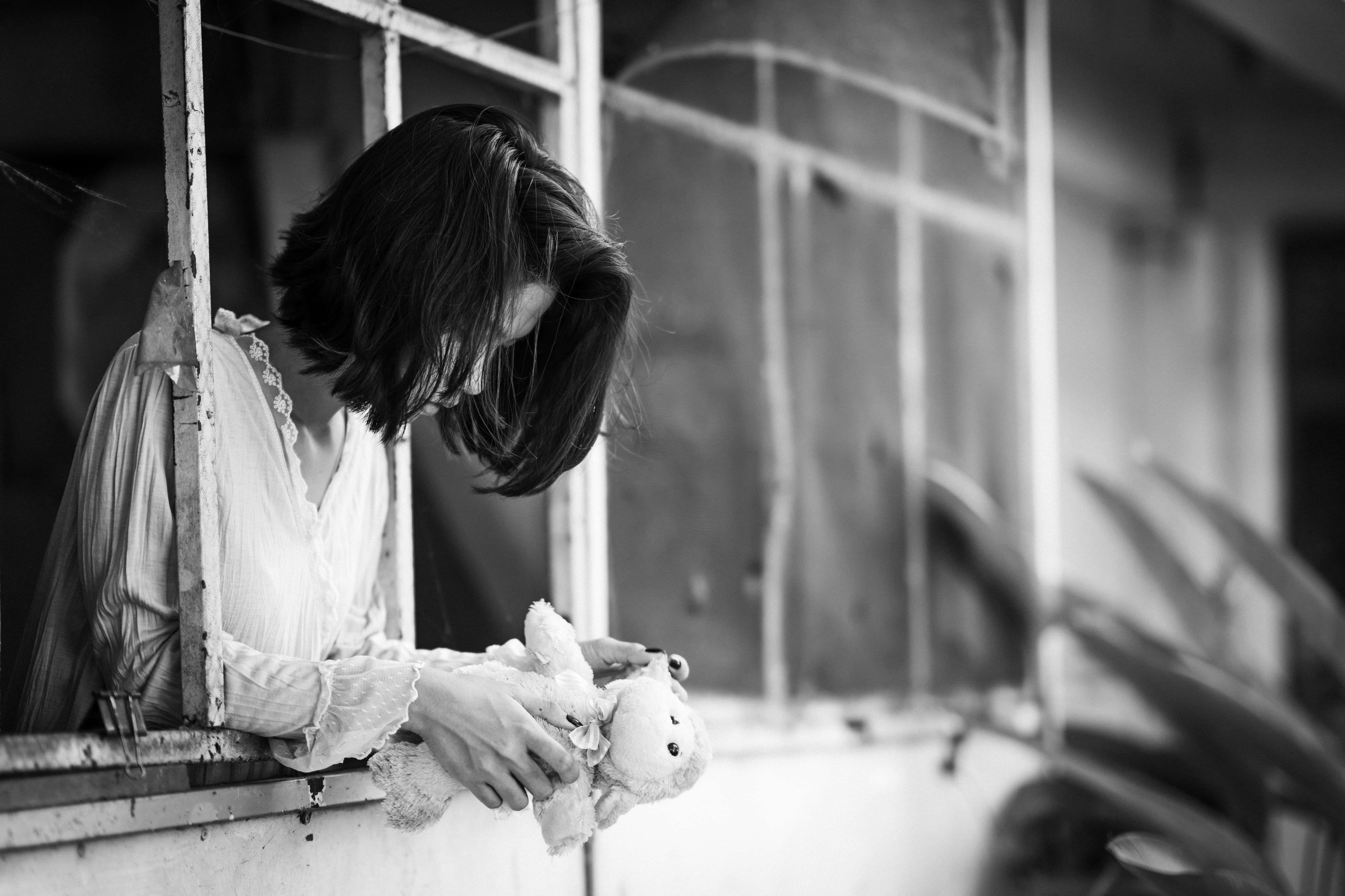 望向窗外的悲傷女孩照片 壁紙 — 一位短黑髮女士從一扇破舊的窗戶探出身子，抱著一隻破舊的泰迪熊，場景為單色調。