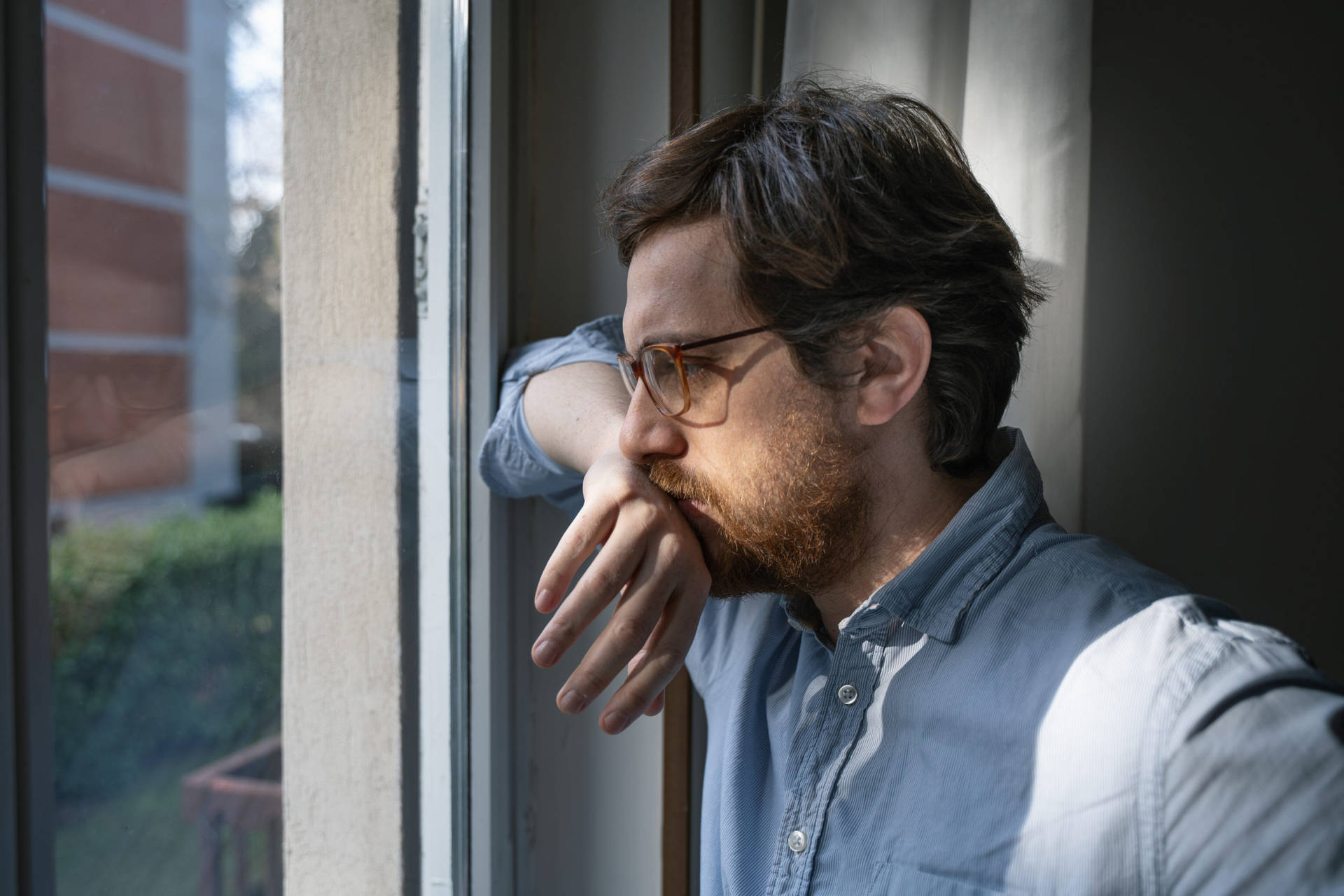 戴眼鏡著藍色恤衫嘅男人，用手托住下巴望出窗外。
