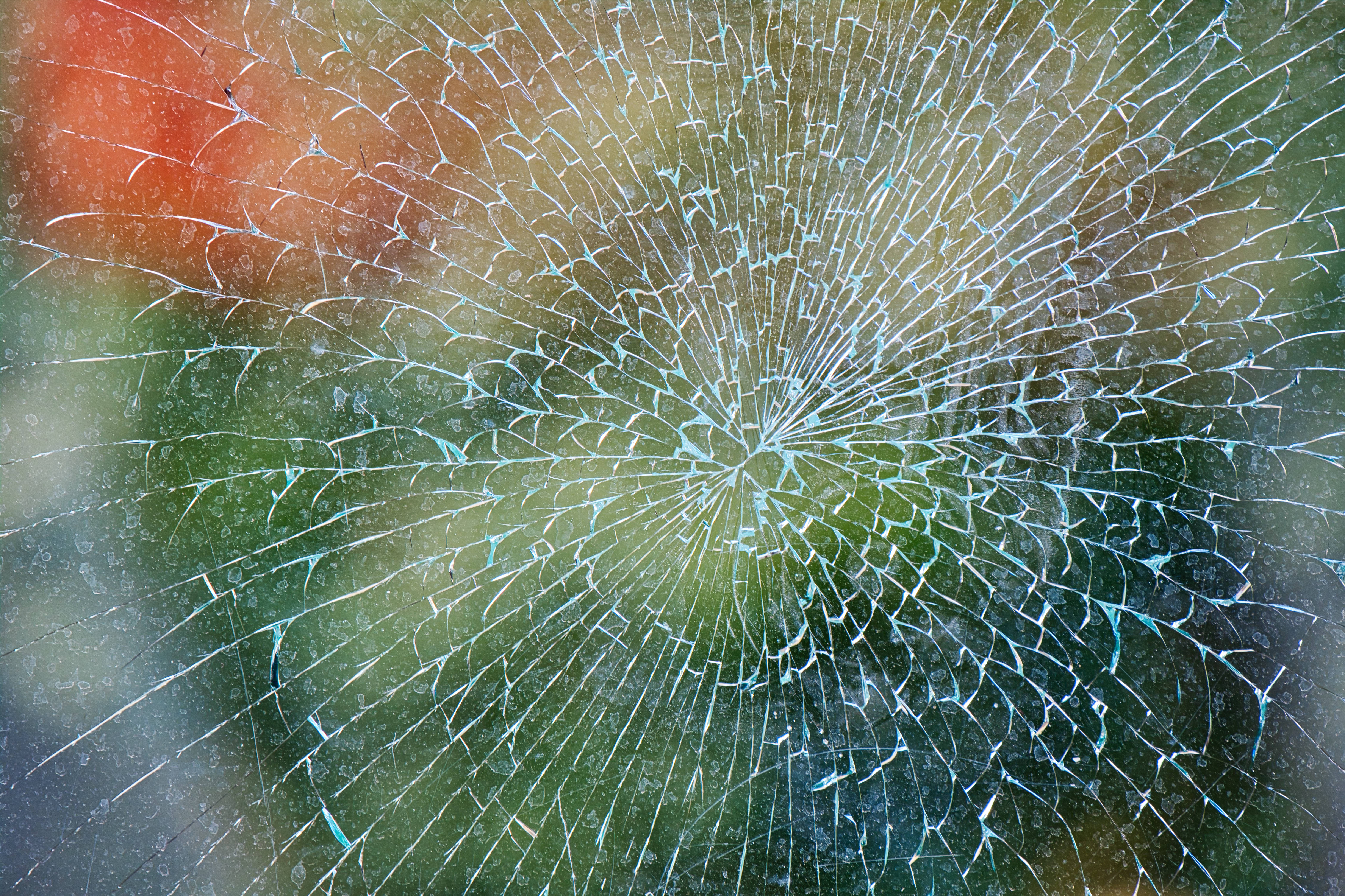 望向綠意的碎窗 壁紙 — 破碎的玻璃表面，從中心撞擊點放射出徑向裂紋，背景為模糊的彩色。