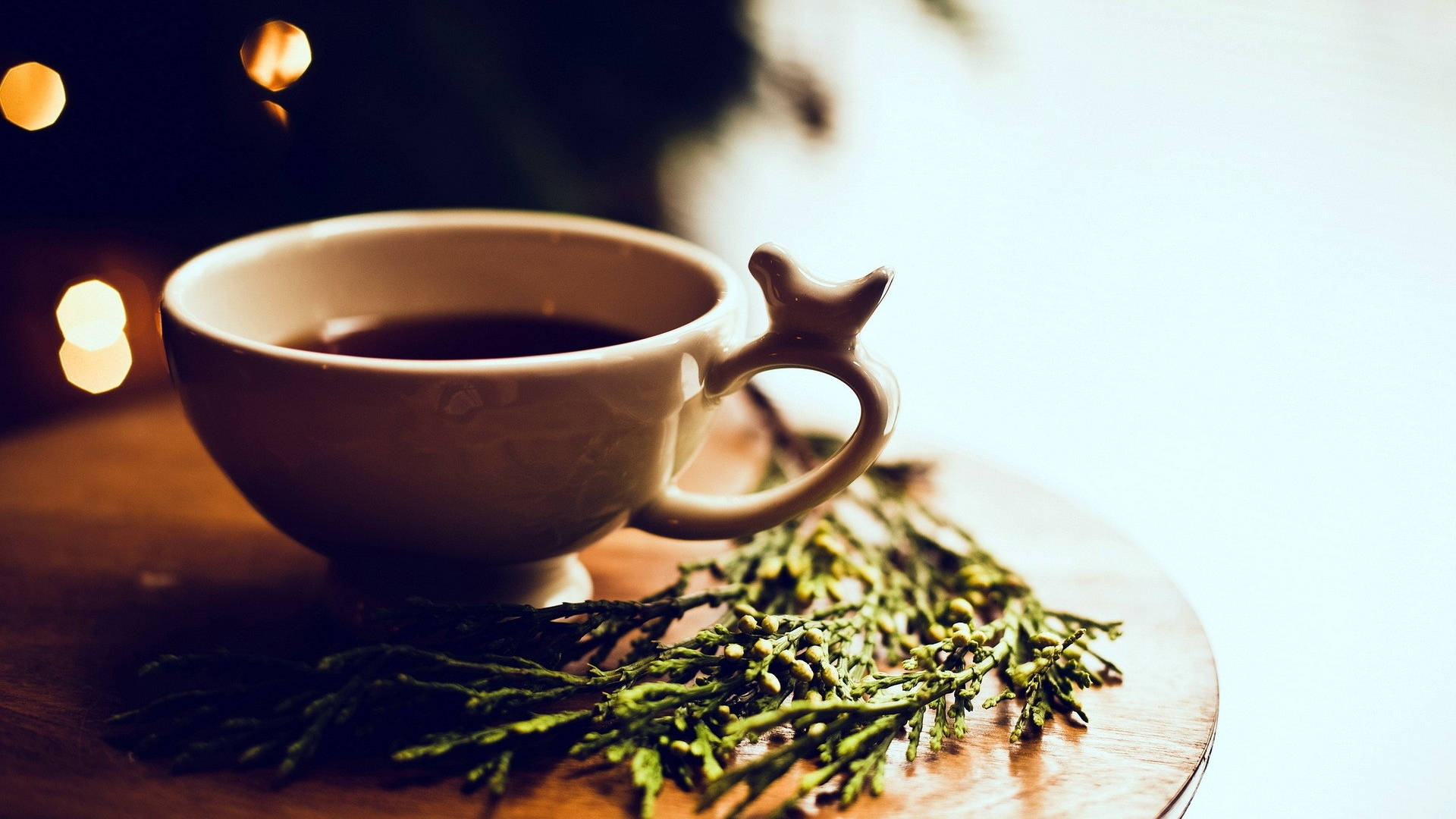 朝顏中的茶杯 壁紙 — 一個帶有星形手柄的陶瓷杯，旁邊是木托盤上的常綠枝條，沐浴在溫暖的散景燈光中。