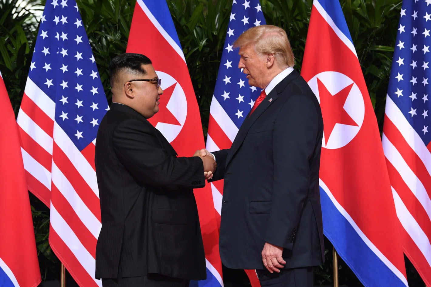 朝鮮領導人金正恩向軍隊敬禮 壁紙 — 兩位世界領袖在交替的美國和北韓國旗前握手，周圍是茂密的綠植。