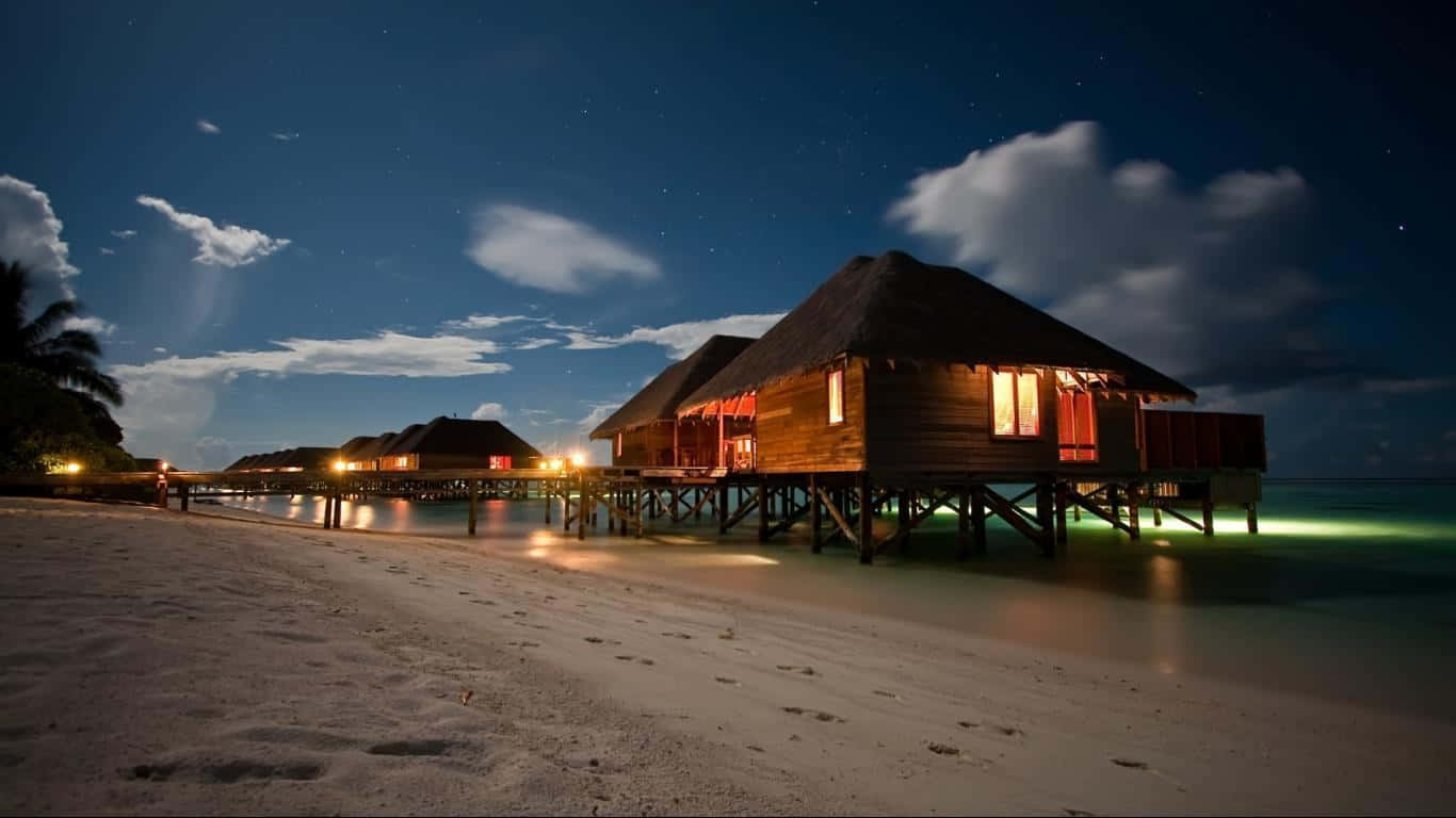 木屋海灘小屋 1366x768 監視器背景 壁紙 — 水上別墅在寧靜海灘上的支柱上溫暖發光，棕櫚樹環繞，星空之夜。