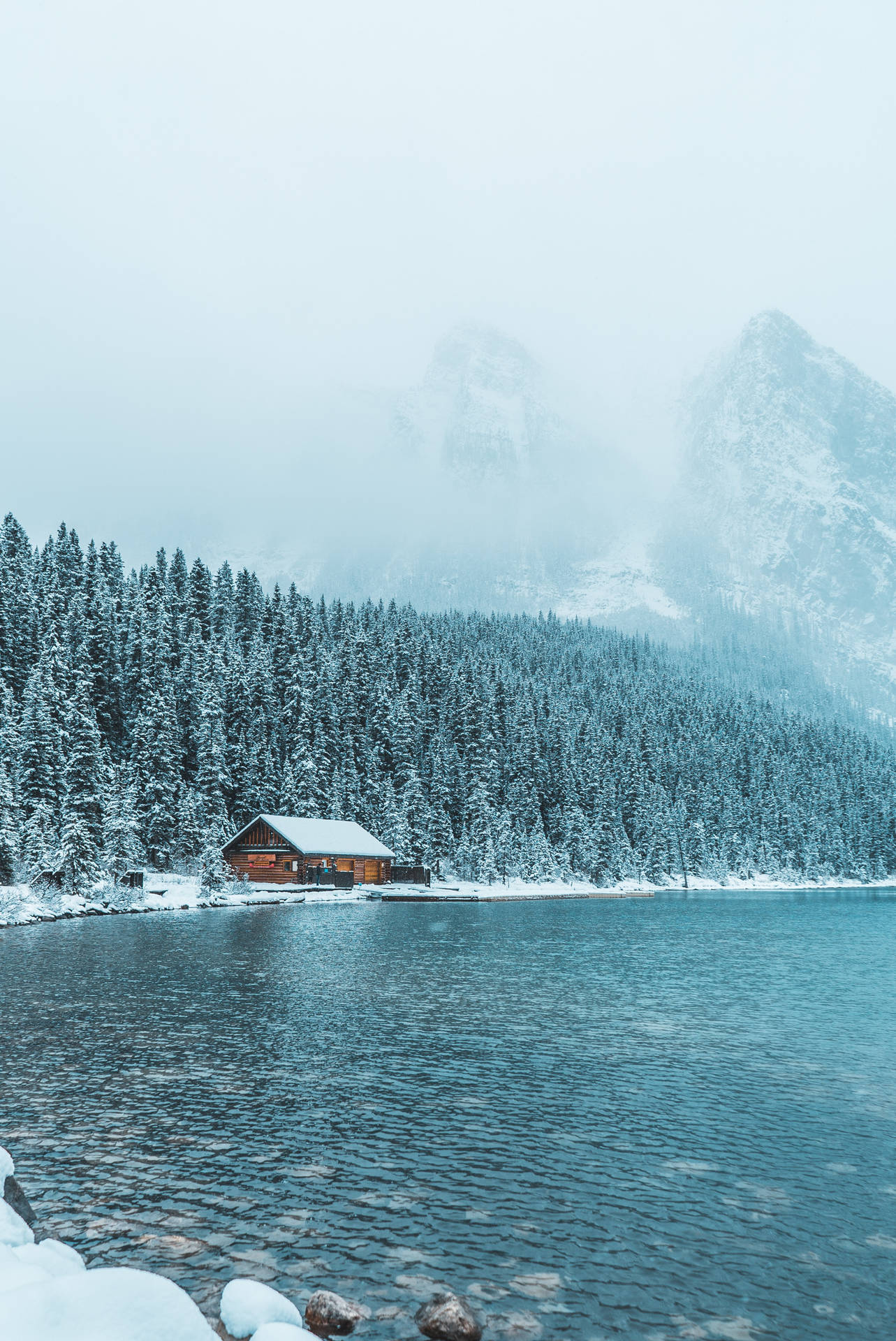 冬季藍色湖邊的雪中小屋，有松樹和霧山。