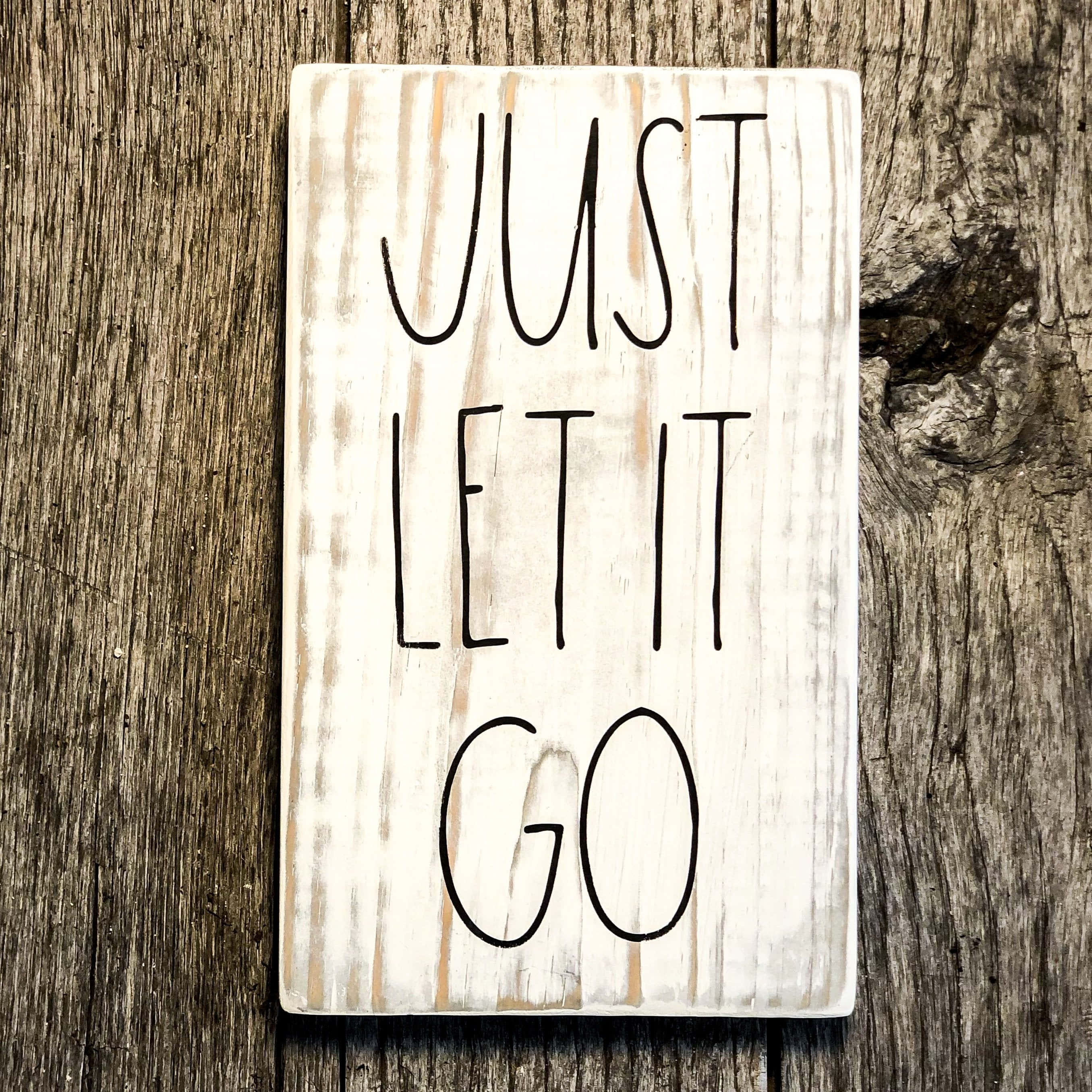 木板上的「JUST LET IT GO」 壁紙 — 一塊經風化的白色木牌，上面有黑色手寫文字「JUST LET IT GO」，擱在粗糙有紋理的木質表面上。