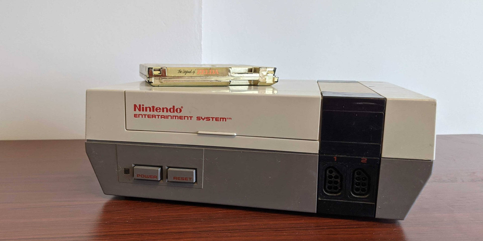 木桌上擺放的NES主機 壁紙 — 米色 Nintendo Entertainment System 主機放在木桌上，上面放著一個金色遊戲卡帶。