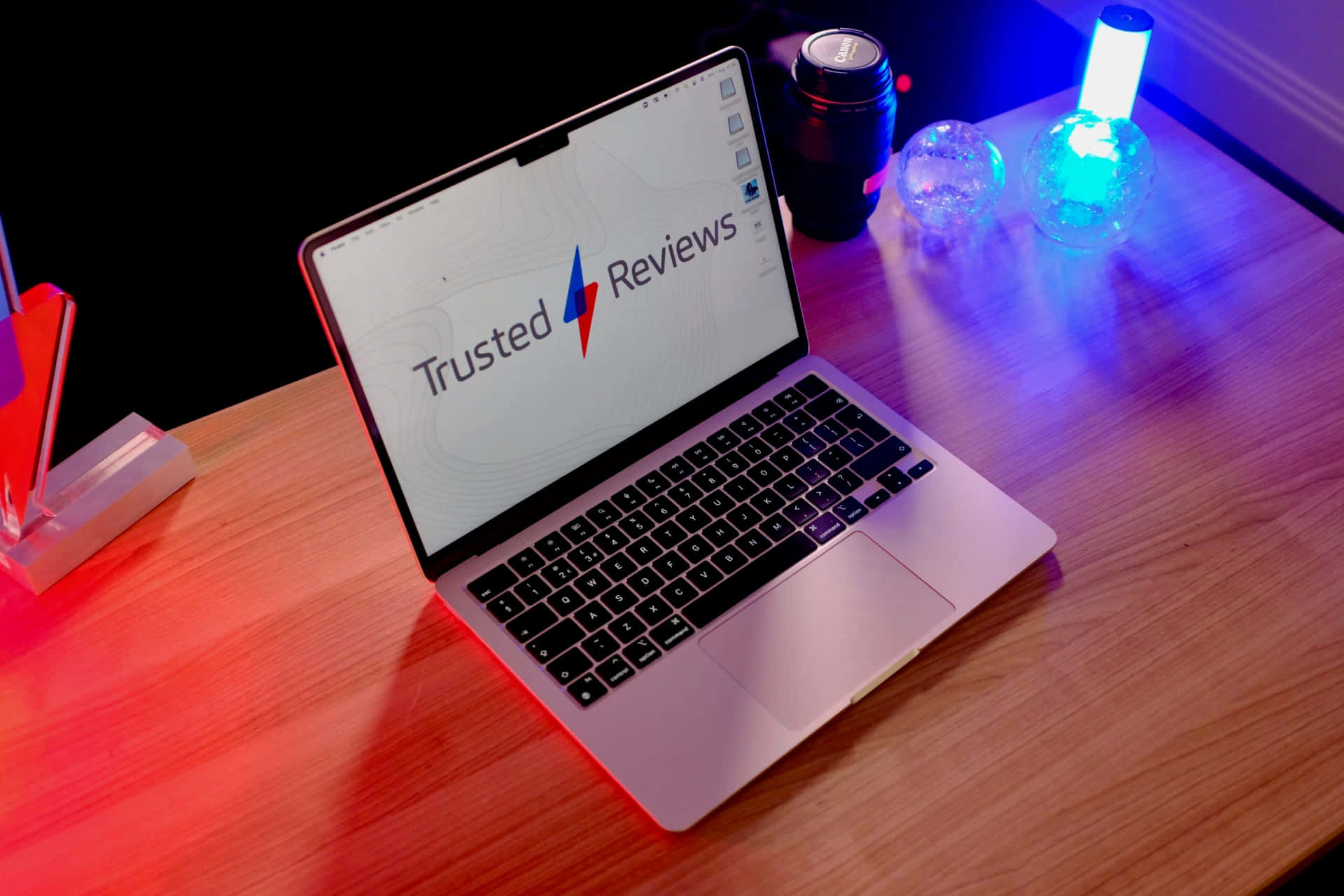 木桌上擺放的銀色 MacBook 壁紙 — 一部銀色手提電腦顯示 Trusted Reviews，放在木桌上，被紅色和藍色霓虹燈裝飾照亮。