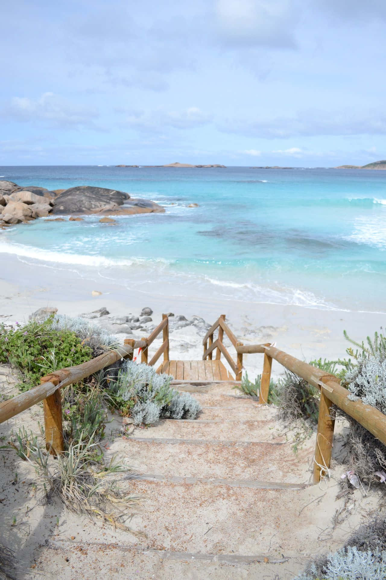 Australia Beach 桌布 壁紙 — 木樓梯穿過沙丘向下延伸至碧綠色的海洋，白色波浪在部分多雲的天空下拍打岩石海岸。