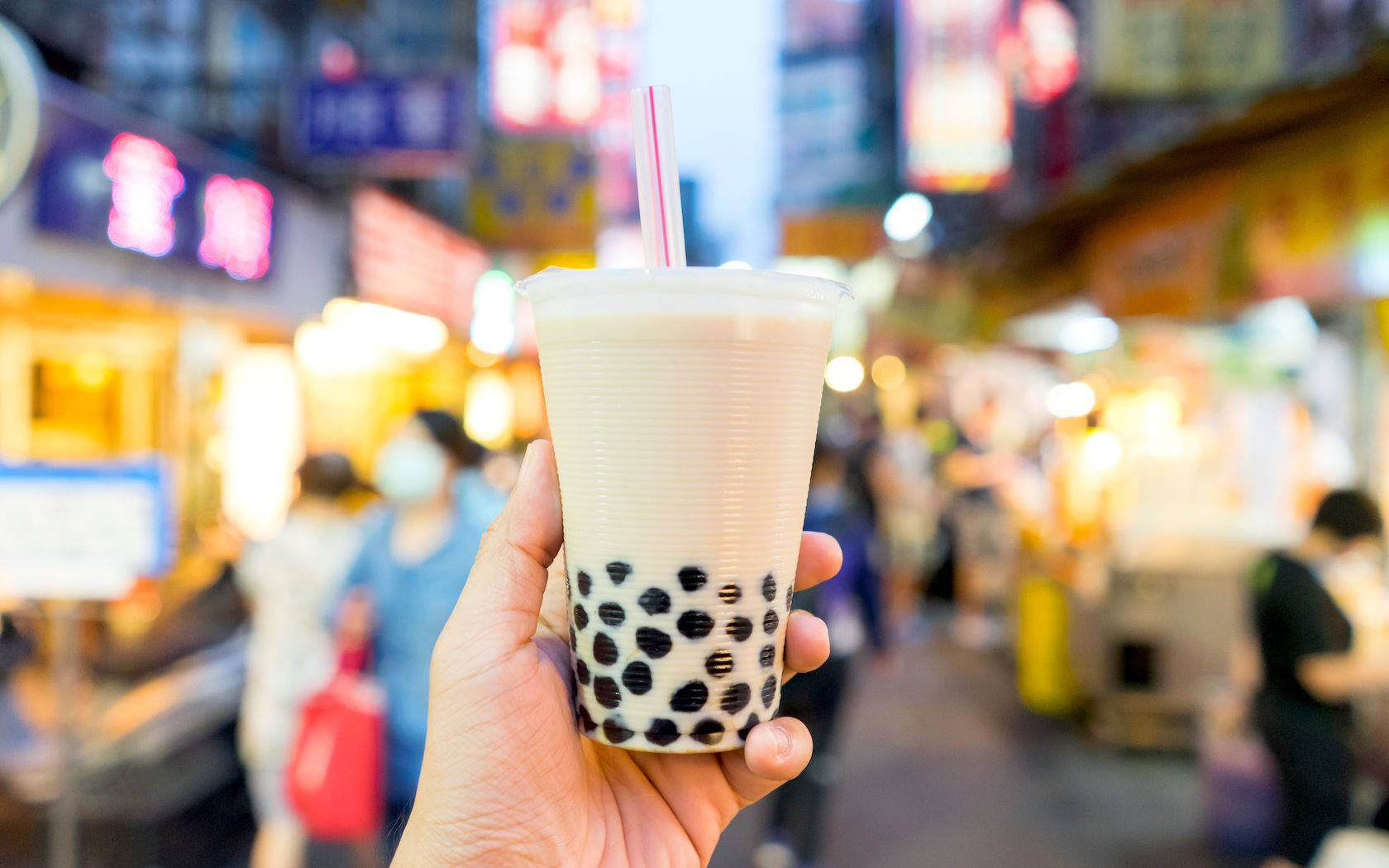 喺繁忙嘅夜市中，手拎住泡泡茶，旁邊有發光嘅招牌。