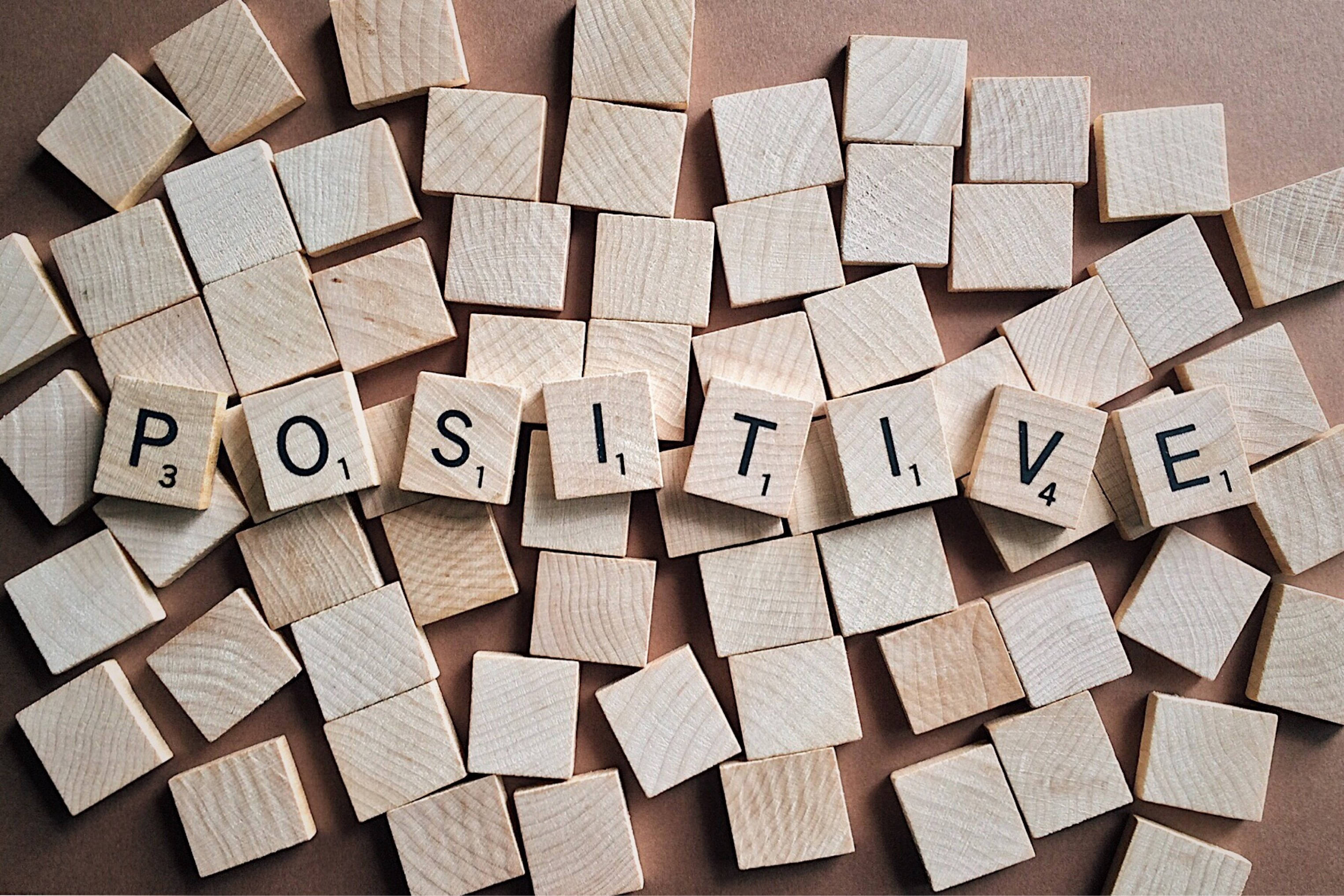 鼓舞人心的 桌布 壁紙 — 木製 Scrabble 字母磚拼出「positive」，周圍散落著空白磚塊，置於溫暖的棕色表面上。