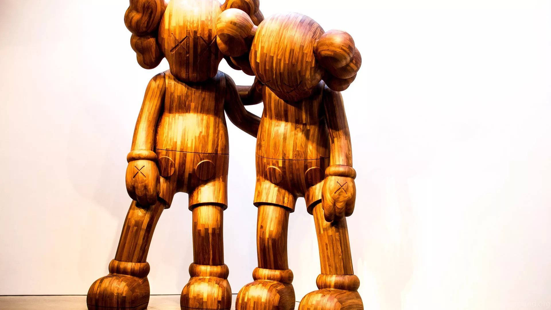 木製KAWS Along The Way 壁紙 — 兩個大型木製KAWS公仔在畫廊環境中，靠著純白牆壁互相擁抱。