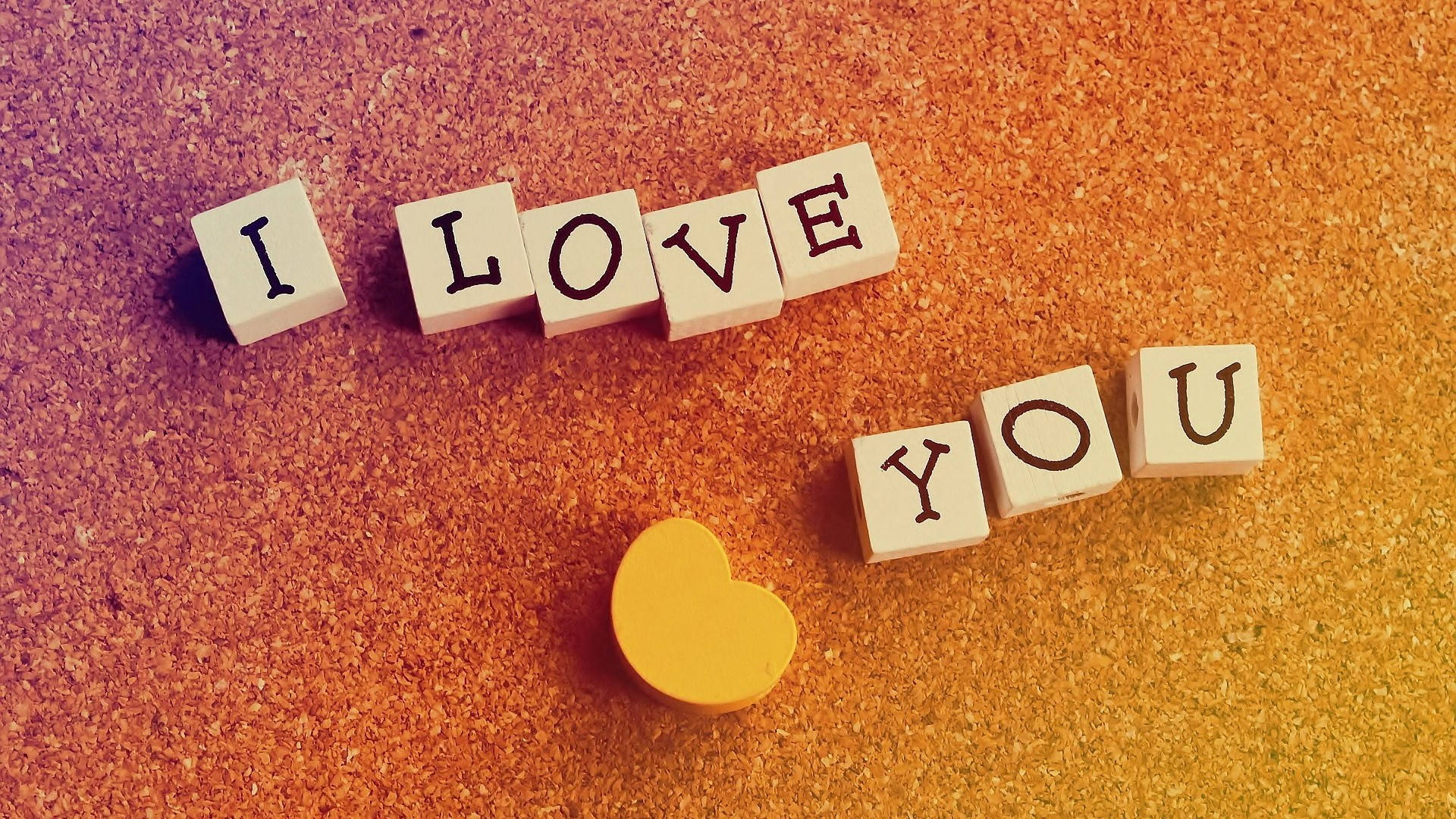 木製字母 我愛你 壁紙 — 木製積木拼出 I LOVE YOU，排列在軟木板上，附近有一個黃色心形，在溫暖漸變燈光下。