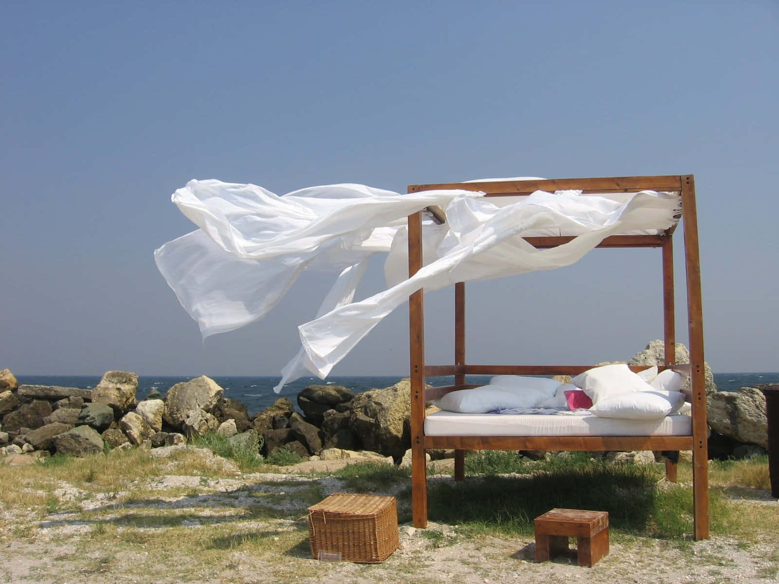 Beach Daybed 桌布 壁紙 — 木製日間床配白色帳篷在風中飄揚，旁邊係岩石海邊，天空晴朗蔚藍。