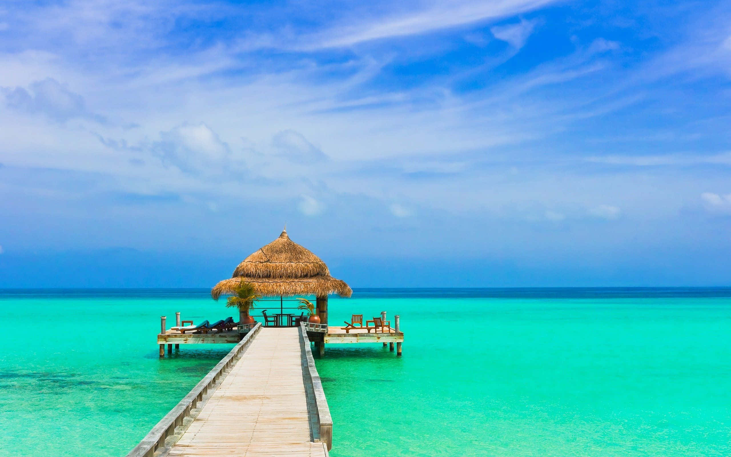 Beach Resort 桌布 壁紙 — 木製碼頭延伸至充滿活力的碧綠海水，通往一座茅草屋頂的水上平房，上方是晴朗的藍天，點綴著縷縷白雲。